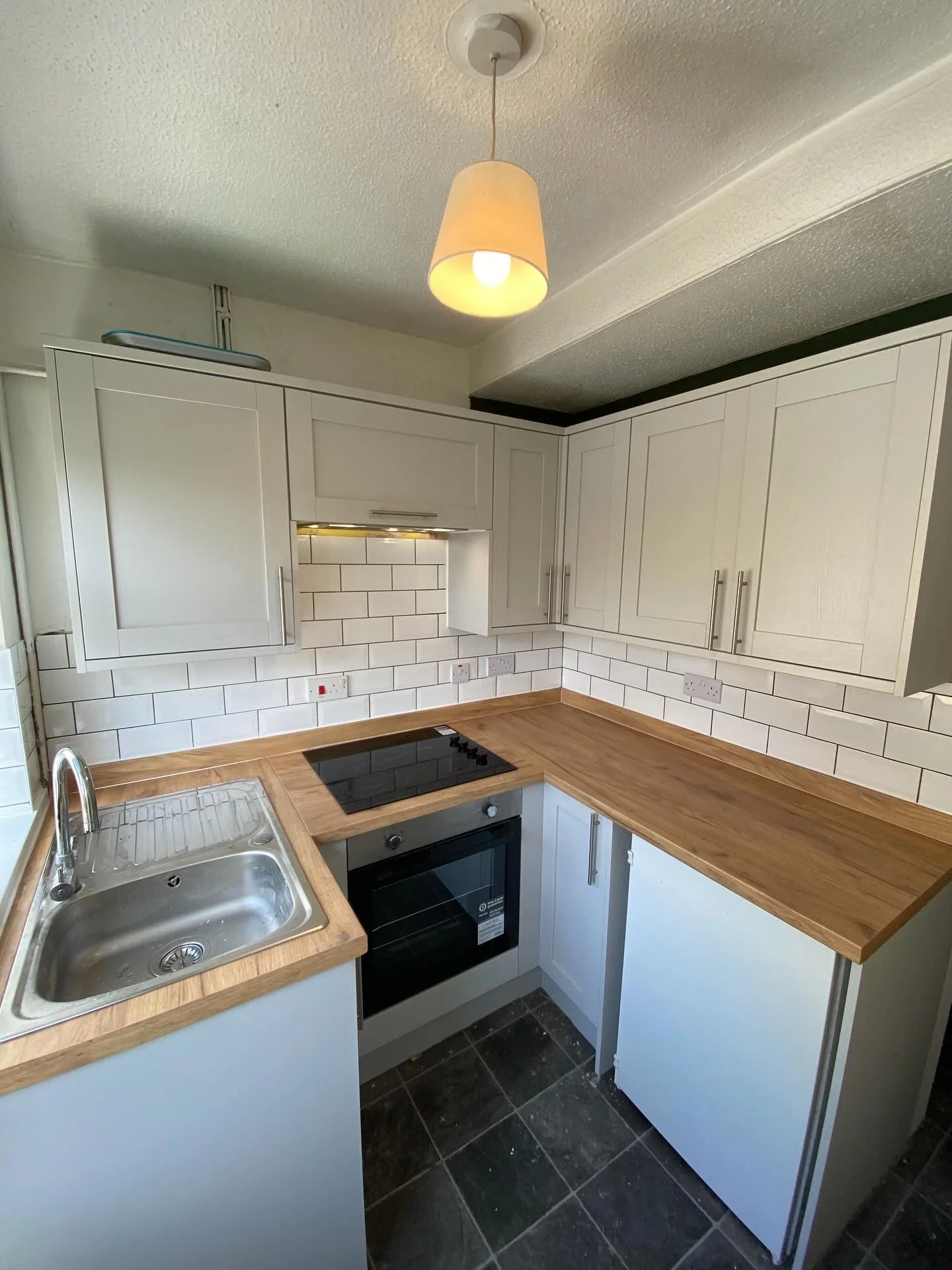 A small kitchen with white cabinets, a wooden countertop, a stainless steel sink, a black electric stovetop, a built-in oven, a tiled backsplash, a ceiling light fixture, and a window with partial natural light.