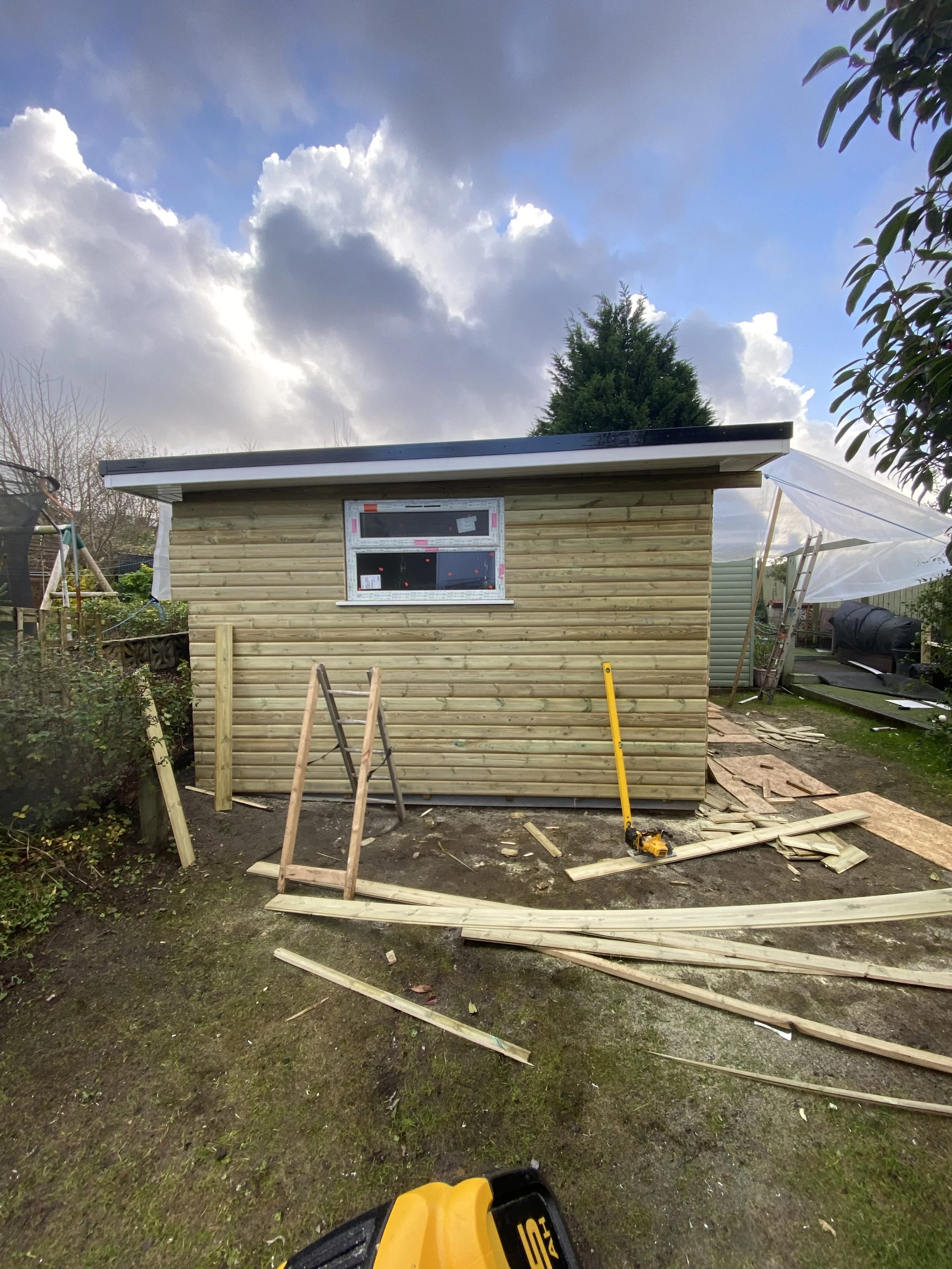 A small wooden shed under construction in a backyard, with building materials and tools around, and a partly cloudy sky above.