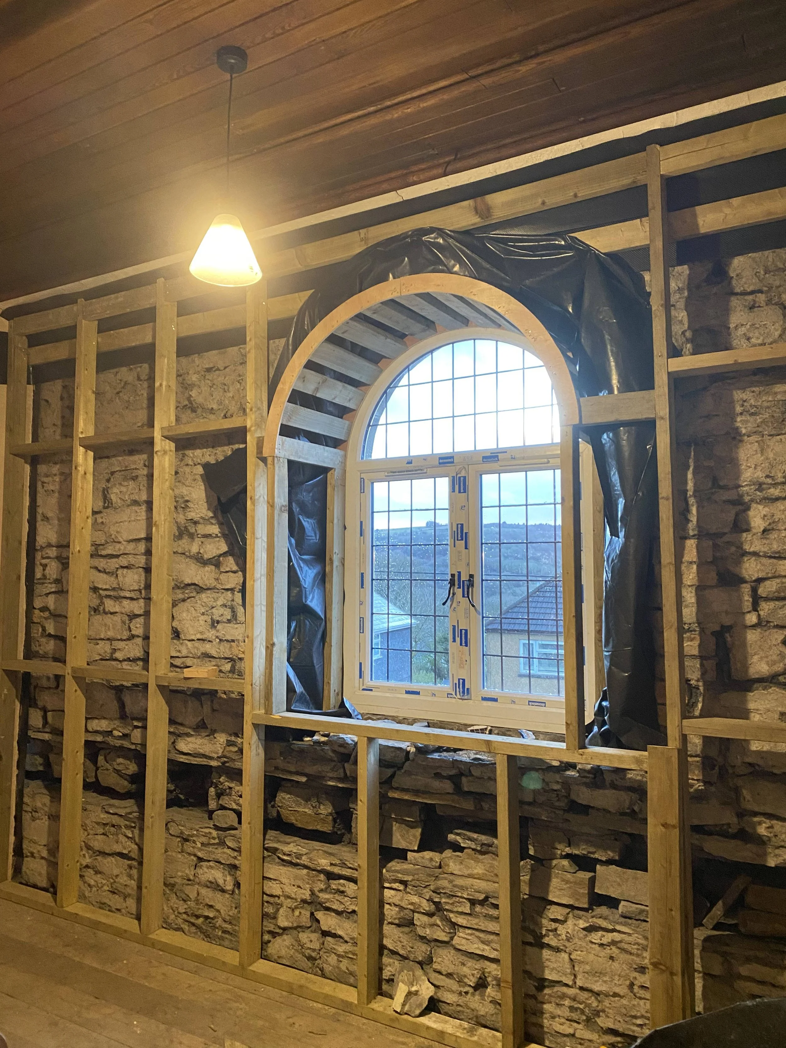 Construction of a stone wall with an arched window, framed with wooden studs, and black waterproofing material around the window opening.
