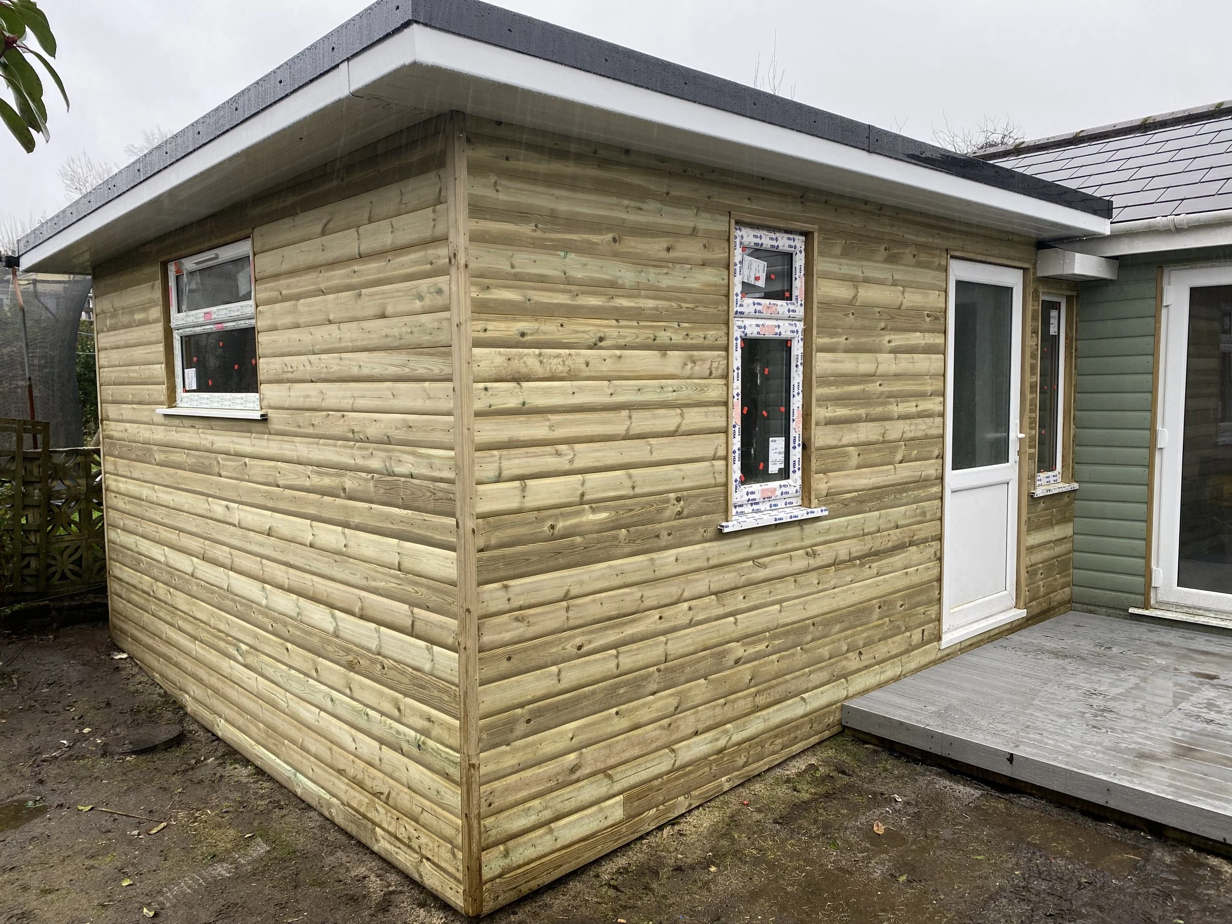 A small wooden shed under construction with three windows and a door, attached to an existing house with green siding, on a cloudy day.
