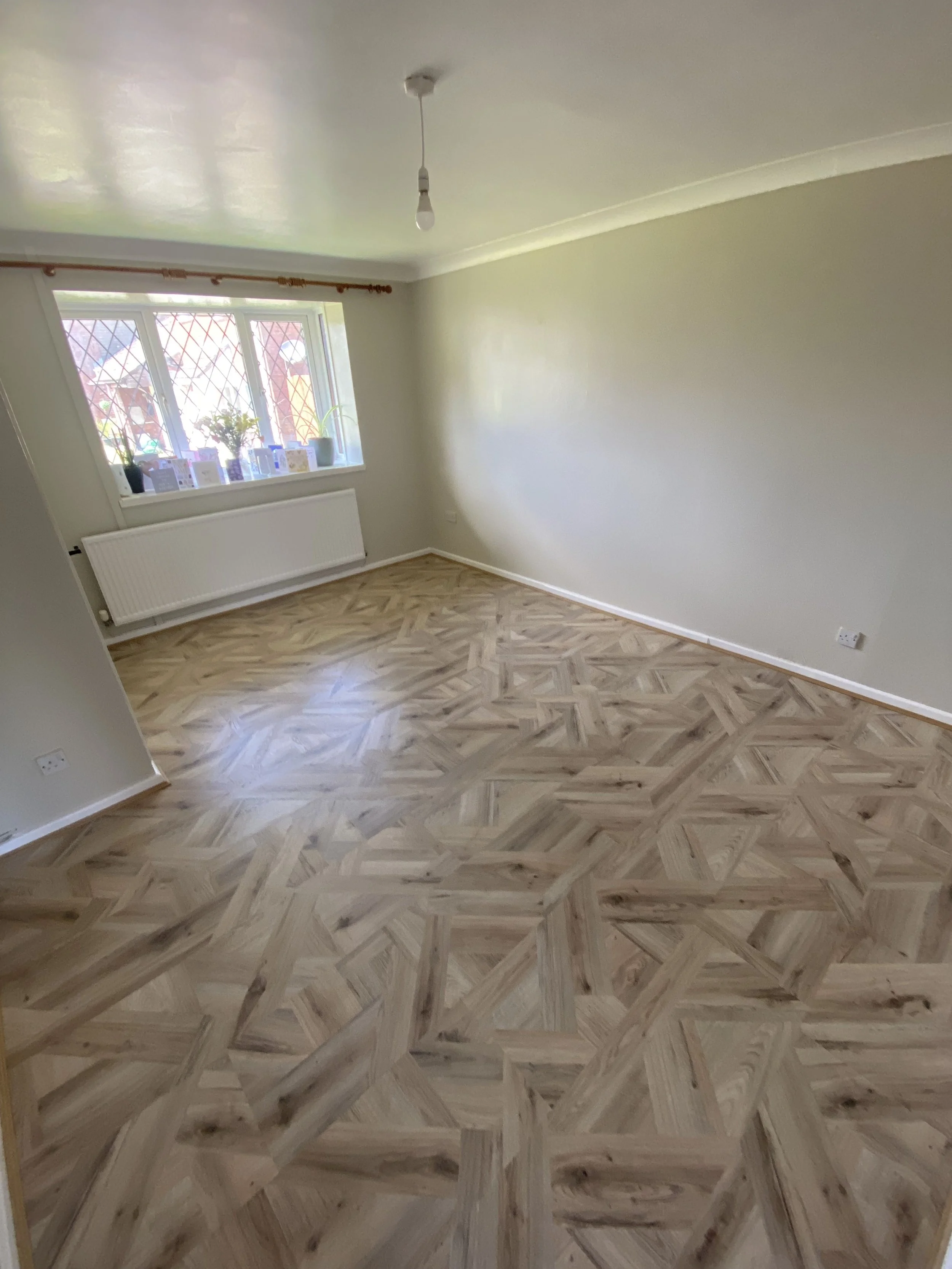 Empty room with light-colored walls, a window with decorative grill, a radiator underneath, and intricate geometric patterned wood flooring.
