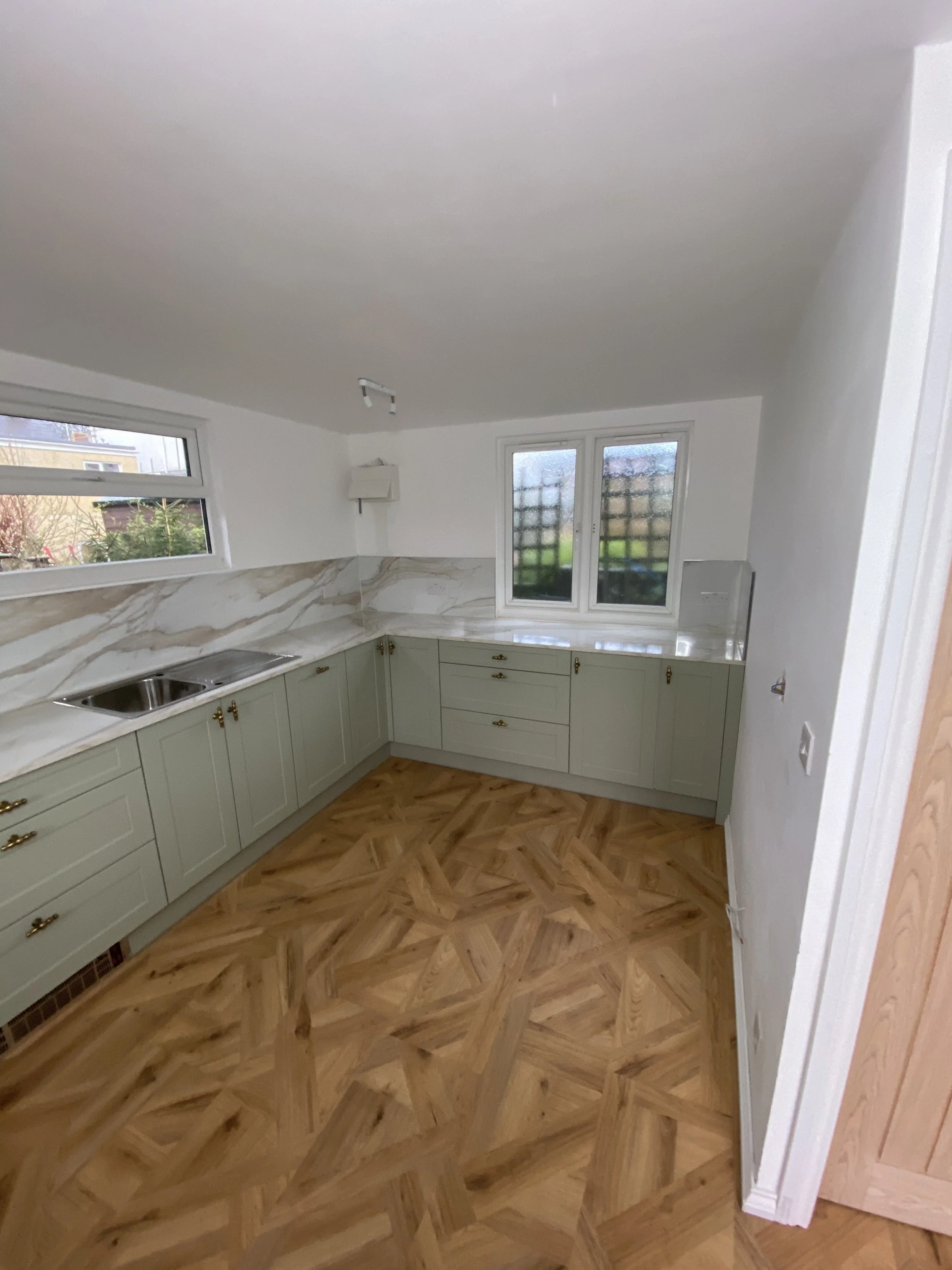 Empty kitchen with light green cabinets, marble backsplash, wooden parquet flooring, and window overlooking a garden.