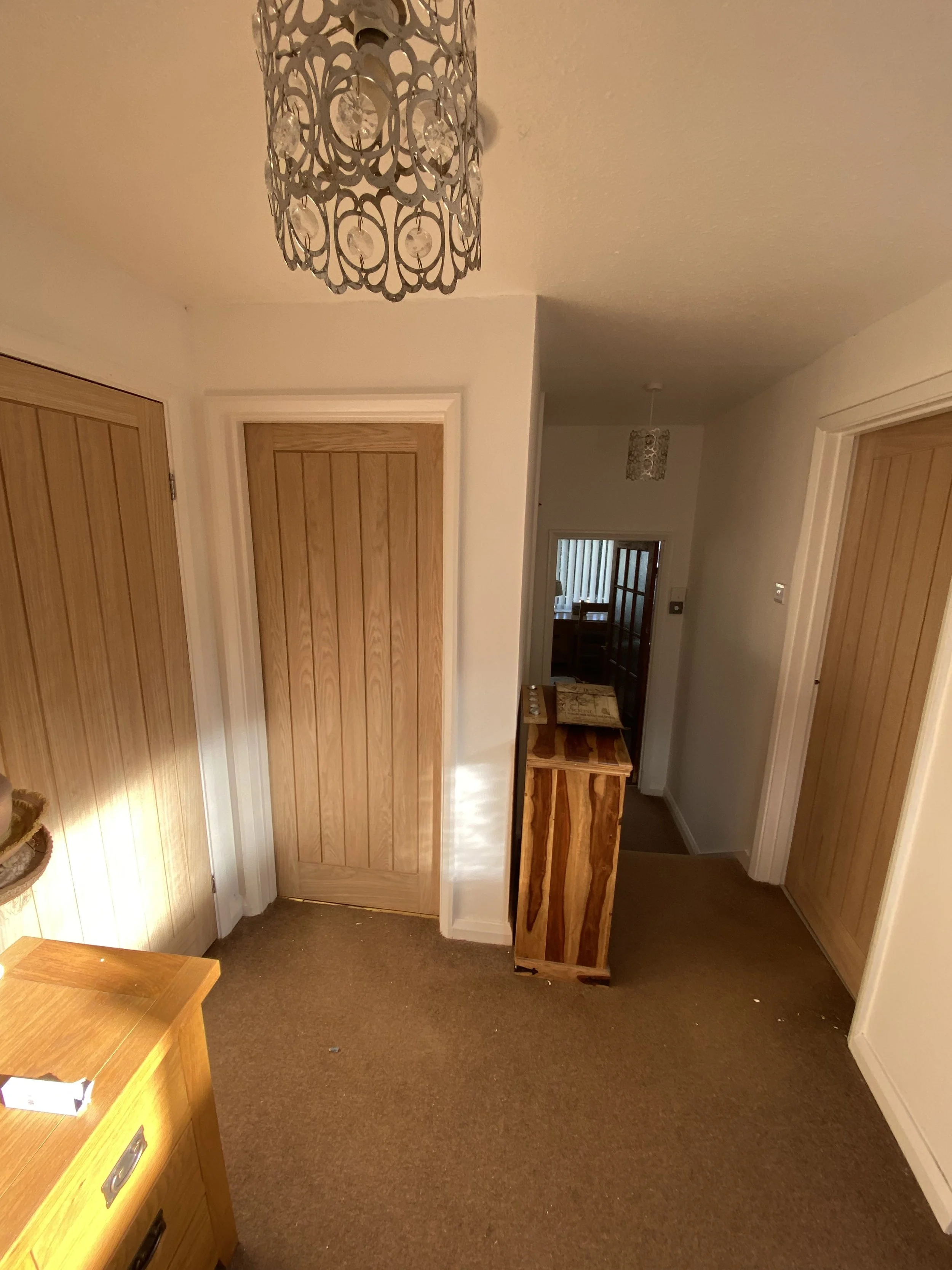 Interior of a room with beige carpet, white walls, and wooden doors. A decorative ceiling light fixture is hanging from the ceiling. There is a small wooden cabinet and a larger piece of furniture in the room.