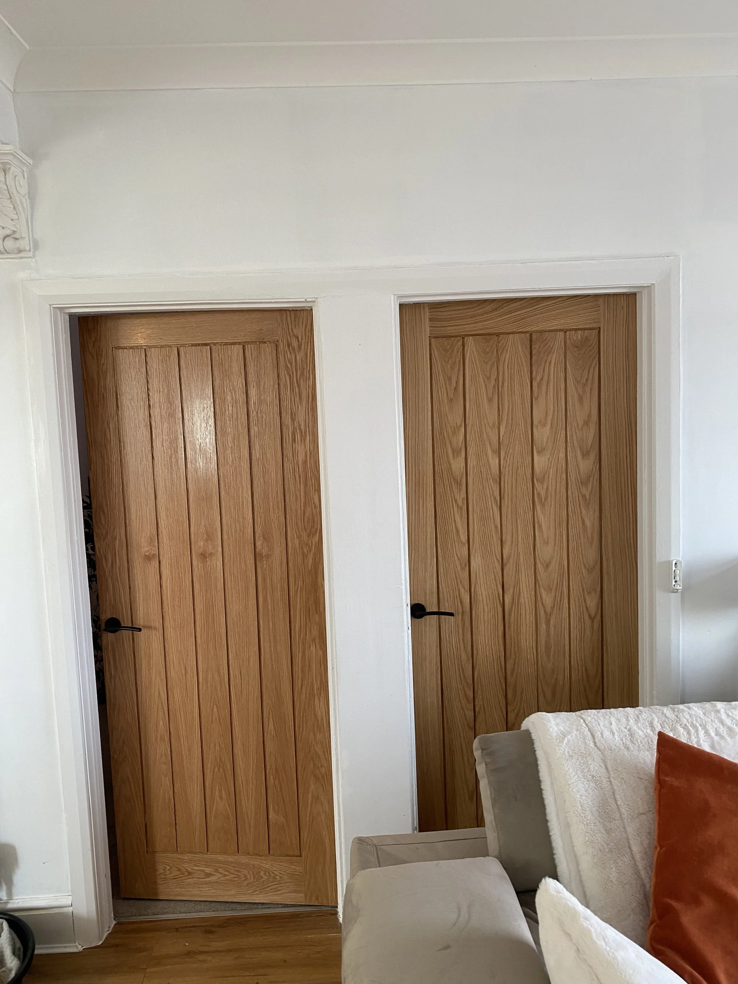 Two closed wooden doors with vertical panels and black handles, set in a white wall, in a living room with a corner of a sofa and a pillow visible in the foreground.