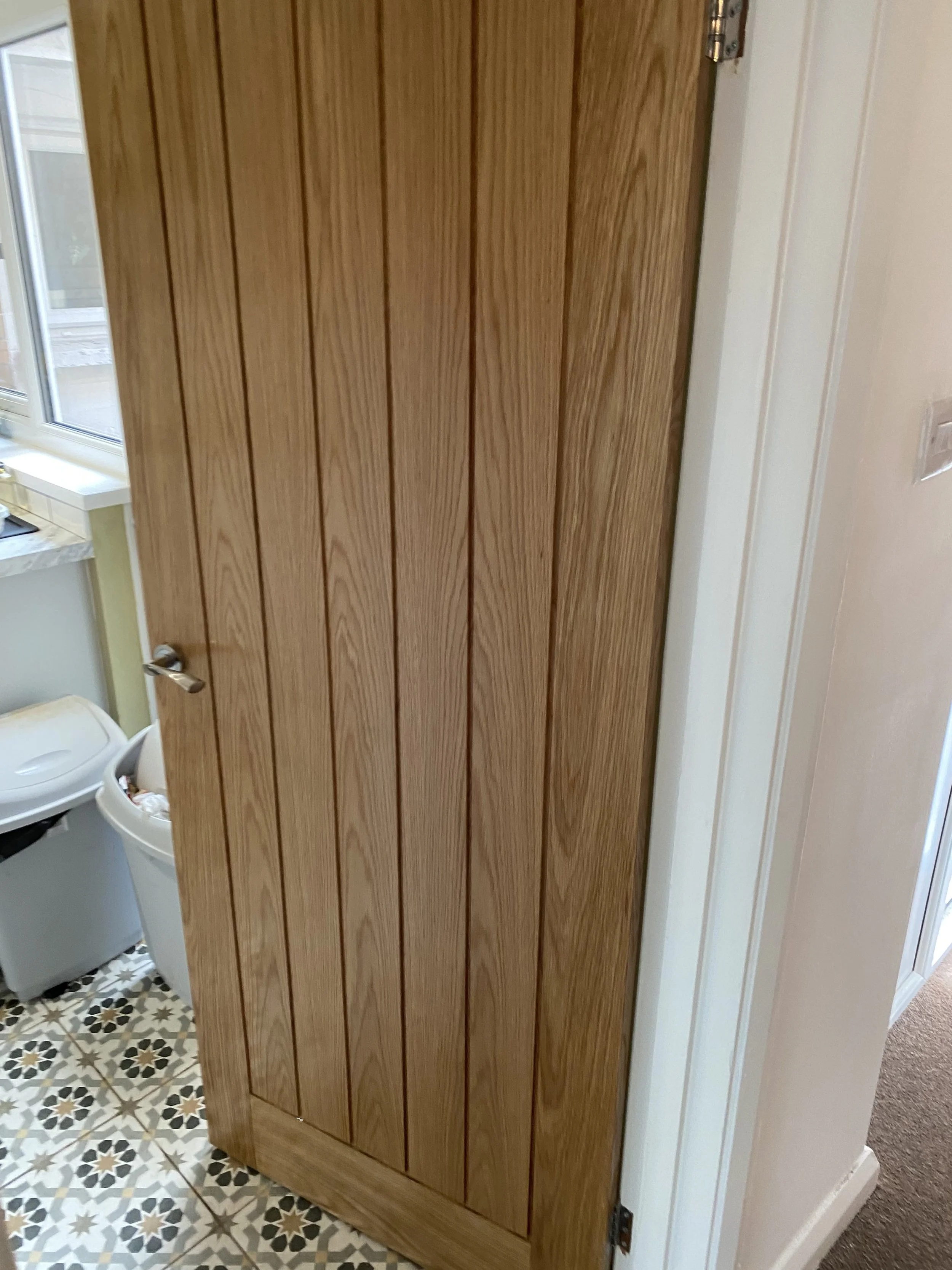 A wooden door in a home interior, partially open, revealing a laundry space with a washing machine and patterned floor tiles.