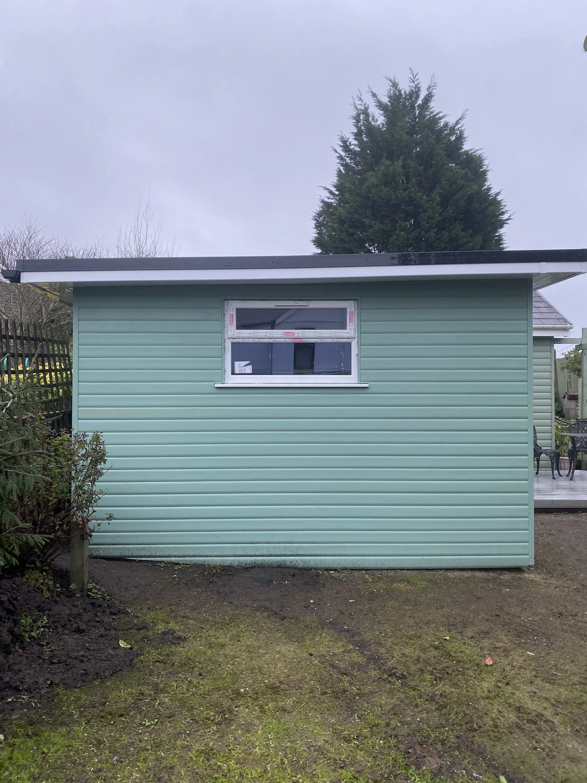 A small, light teal-colored utility building or shed with a single rectangular window, located outdoors in a backyard with a patchy grassy area and a large tree in the background.
