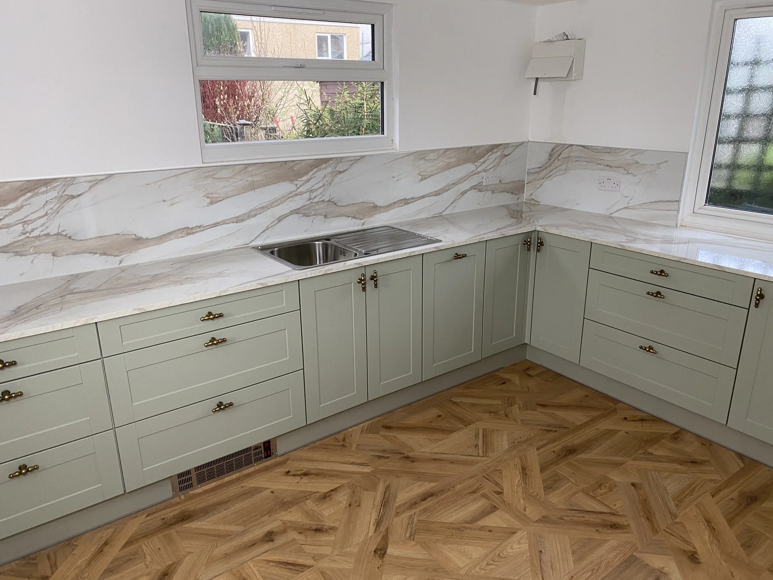 Kitchen with cream-colored cabinets, marble backsplash, and hardwood floor in an L-shaped layout, with windows showing outside greenery.