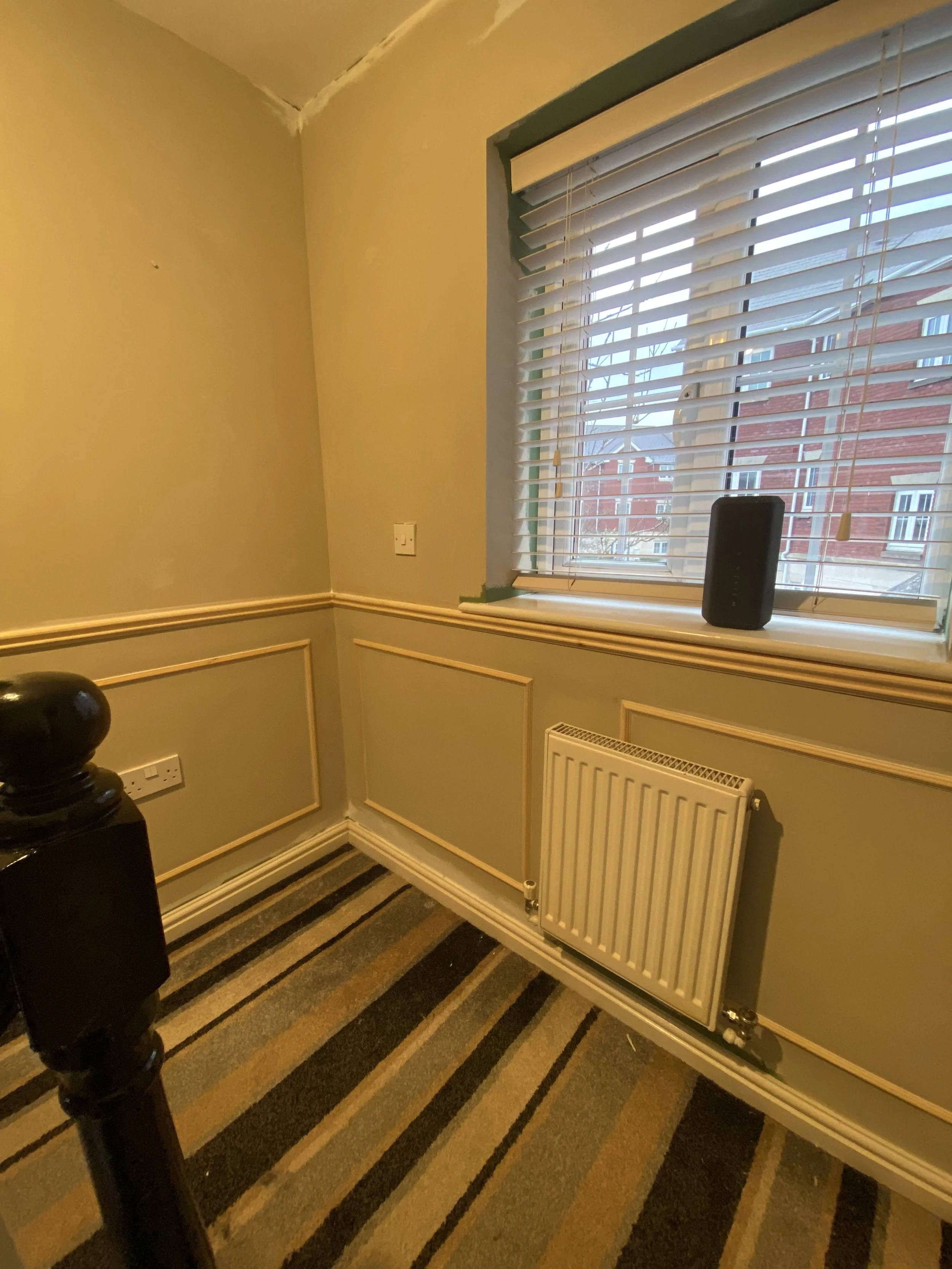 A corner of a room with light green walls and cream-colored wainscoting, a striped carpet in shades of black, gray, and yellow, a large window with closed blinds, and a black speaker on the windowsill.