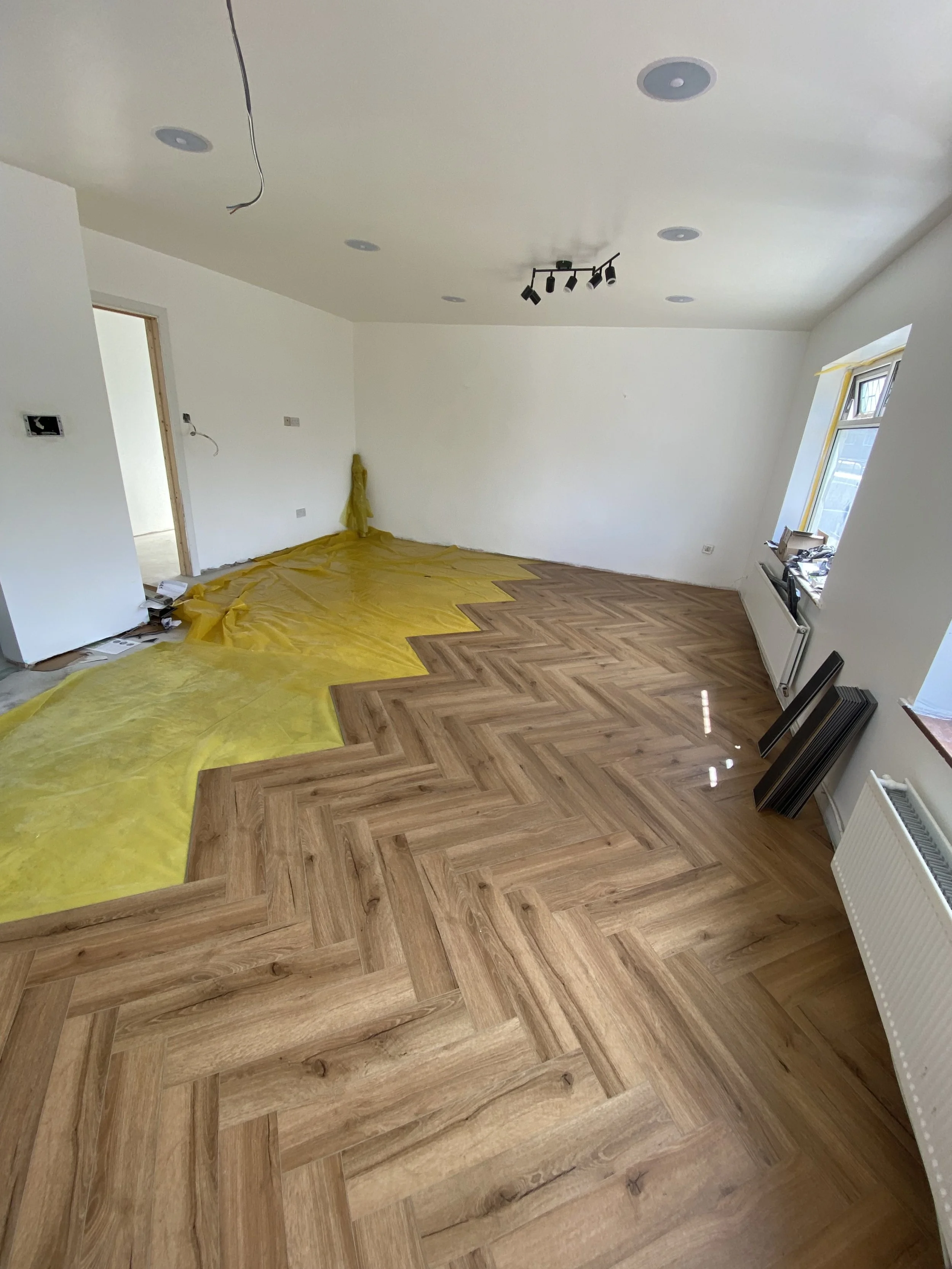 Room under renovation with partially installed wood herringbone pattern flooring, yellow protective covering on part of the floor, and a window letting in natural light.