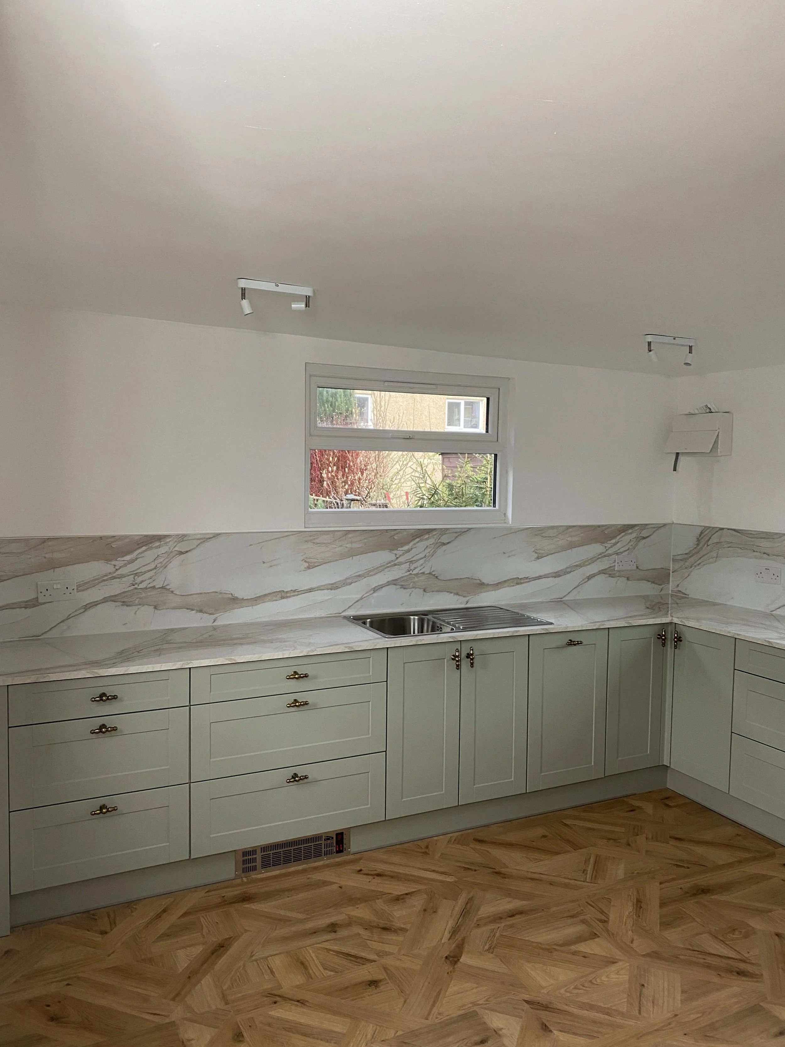 Kitchen with light green cabinets, marble backsplash, stainless steel sink, and small window above the countertop. Wooden parquet flooring and white walls.