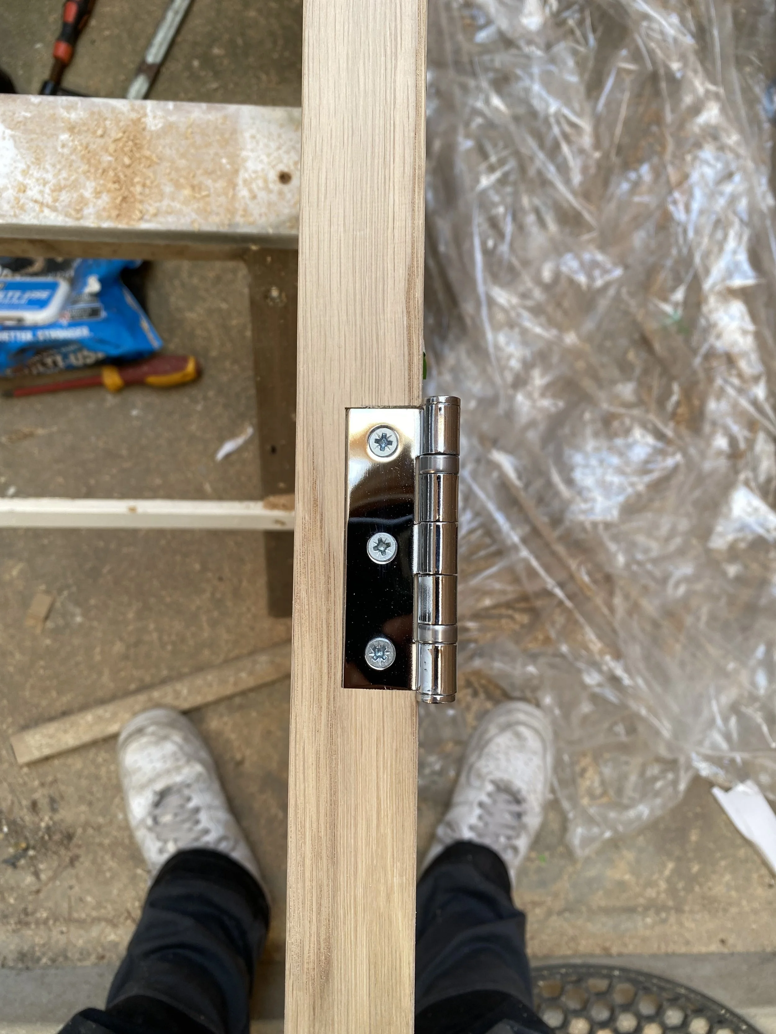 Close-up of a hinge attached to a wooden surface during construction, with tools and materials in the background.