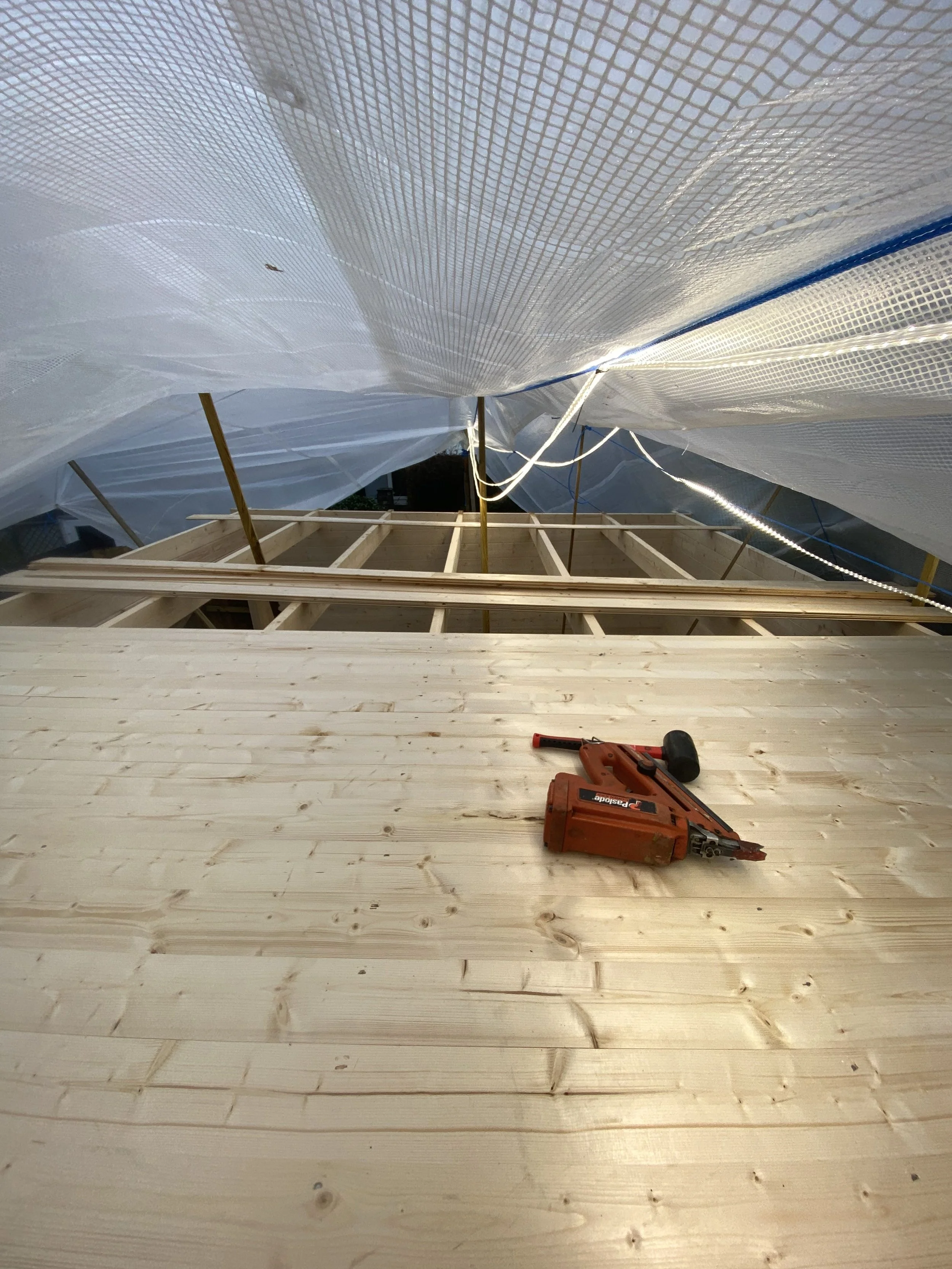 View of a construction site with a wooden structure in progress, a wooden floor, and construction tools including a stapler. Plastic sheeting covers the scaffolding and roof support.