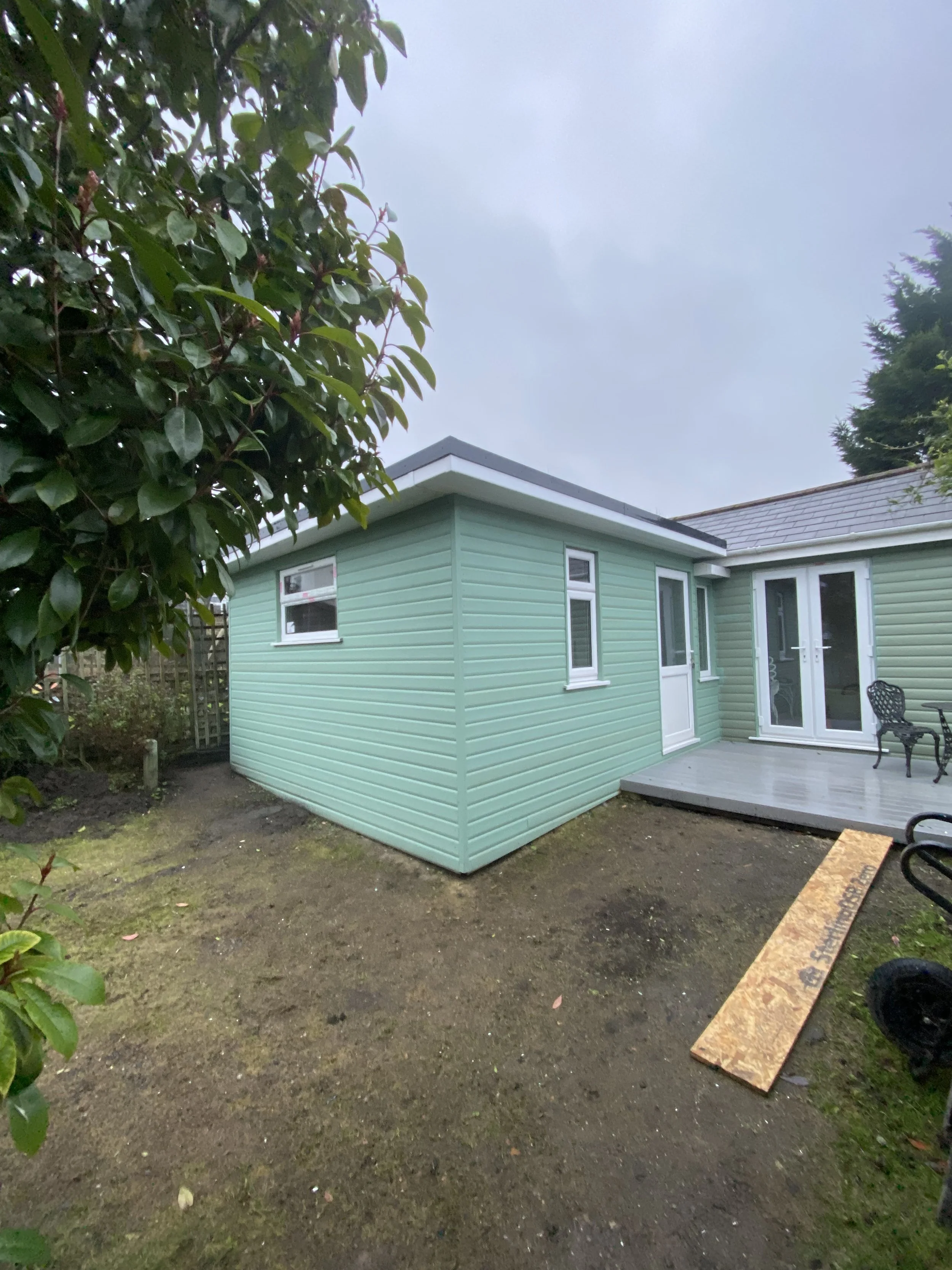 A mint green house with white window frames and a small porch with black chairs, outside on a cloudy day.
