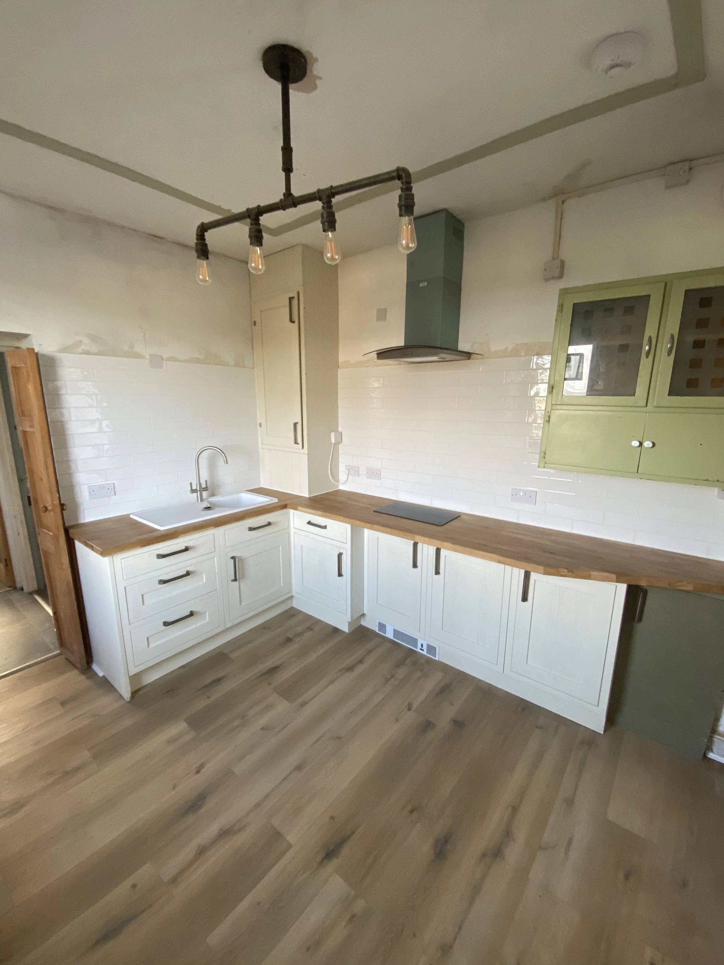 A modern kitchen with white cabinets, wooden countertops, and a green wall-mounted cabinet. The kitchen has a black induction cooktop, a sink with a chrome faucet, and a black industrial-style light fixture with exposed bulbs hanging from the ceiling