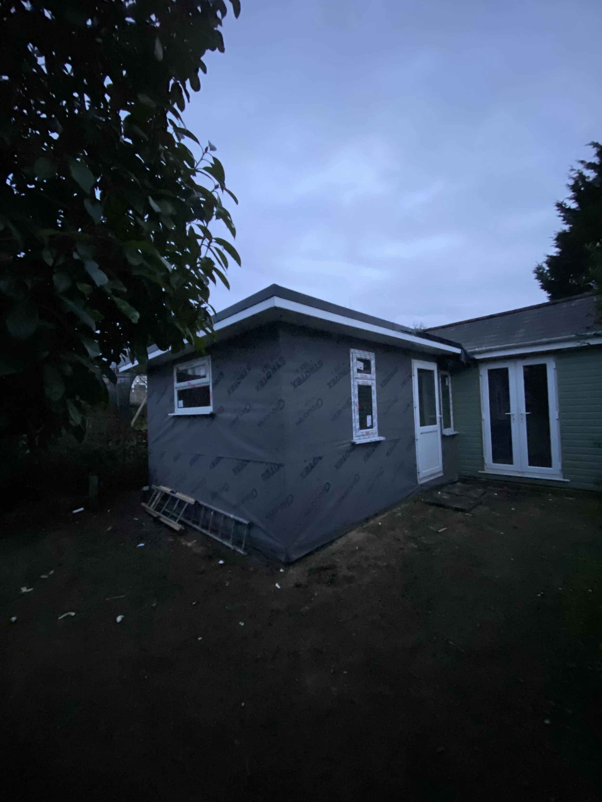 A small building under construction or renovation, with gray weather-resistant sheathing on the exterior, a ladder resting against it, and a partly cloudy evening sky above.