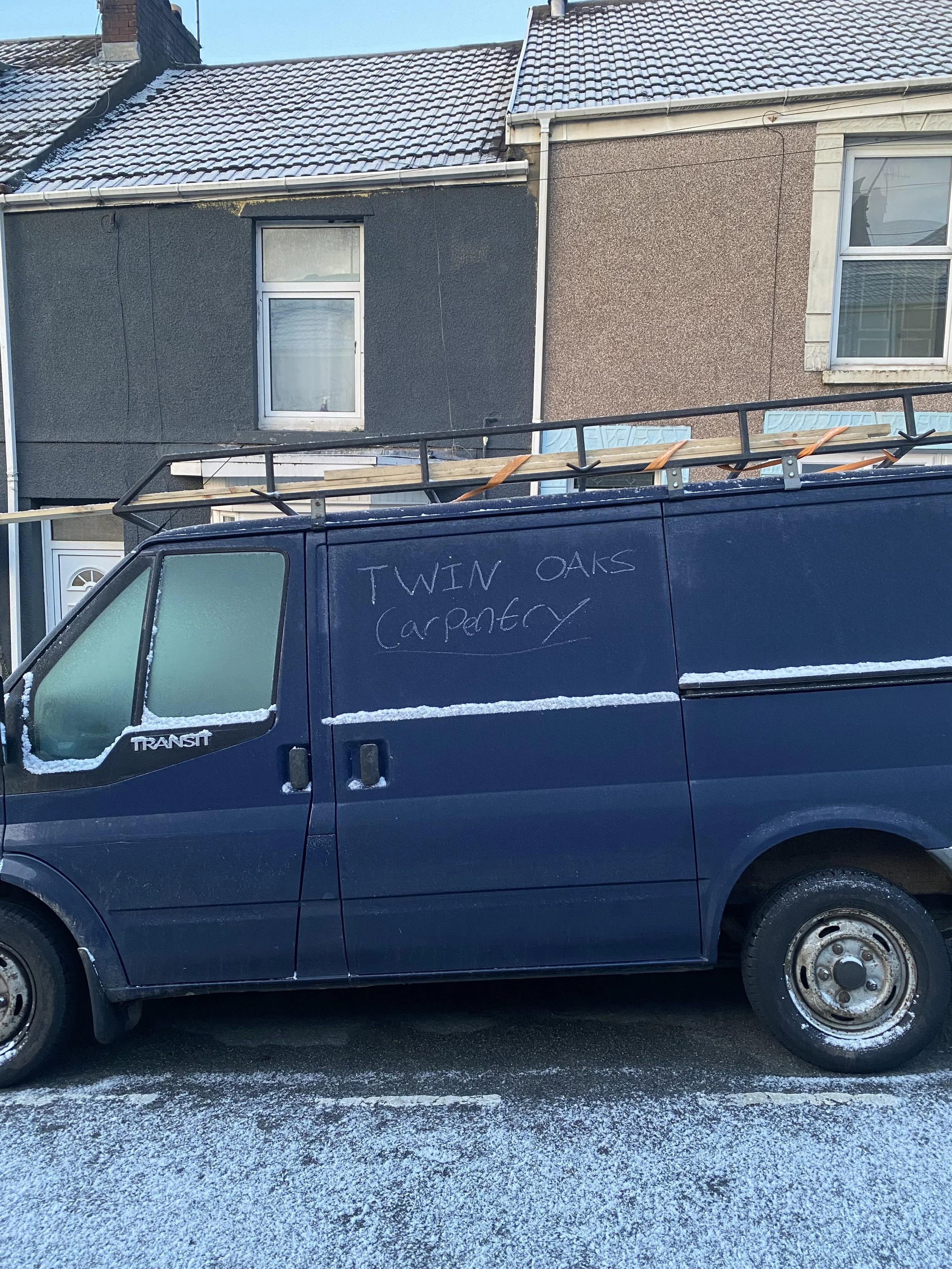 A blue work van parked on a snowy street with two houses in the background. The van has a ladder on top and the words "TWIN OAKS carpentry" written on the side.