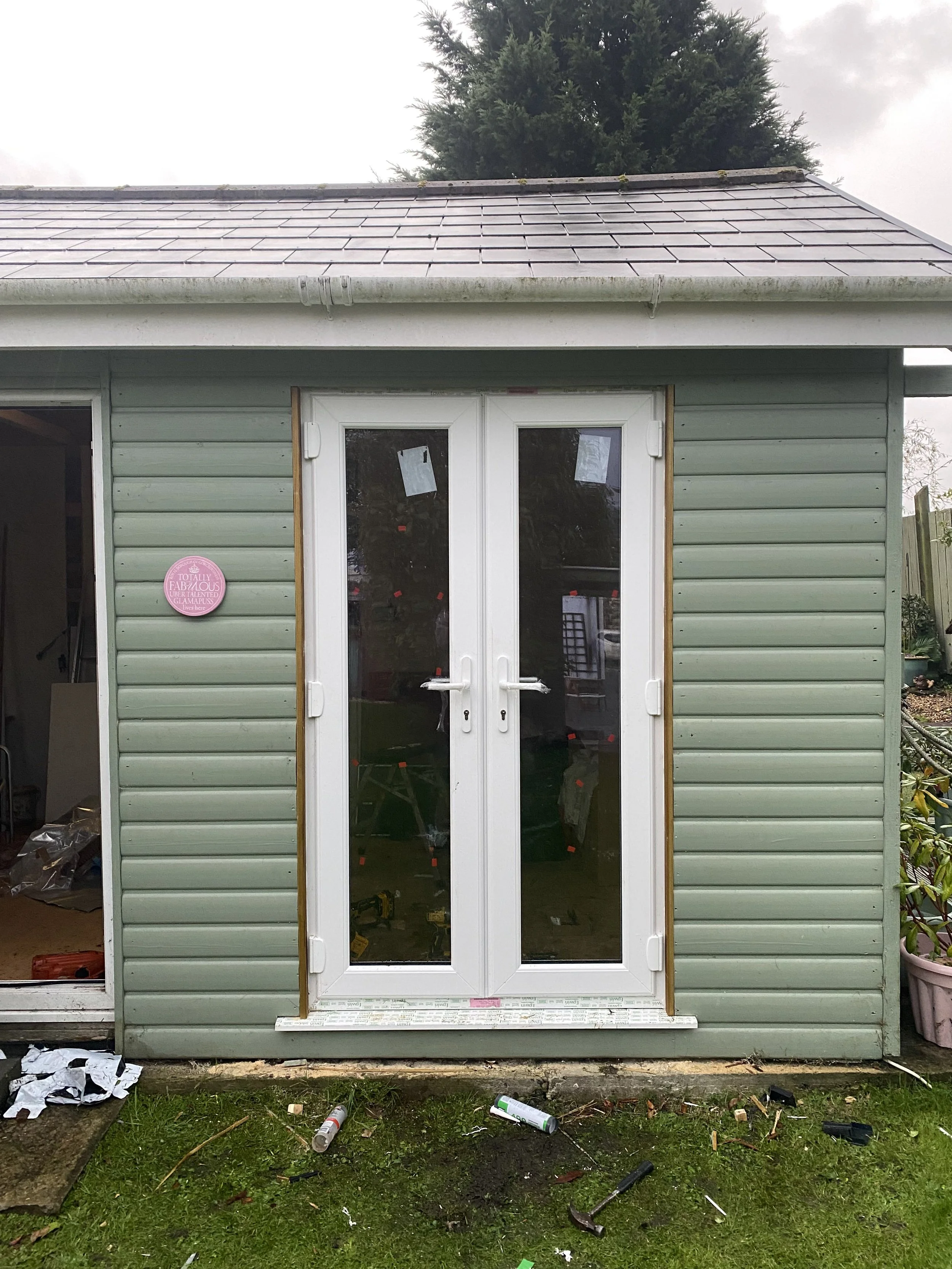 A small green shed with a gray shingled roof and a set of white double glass doors, surrounded by tools and construction materials, indicating ongoing renovation or construction.