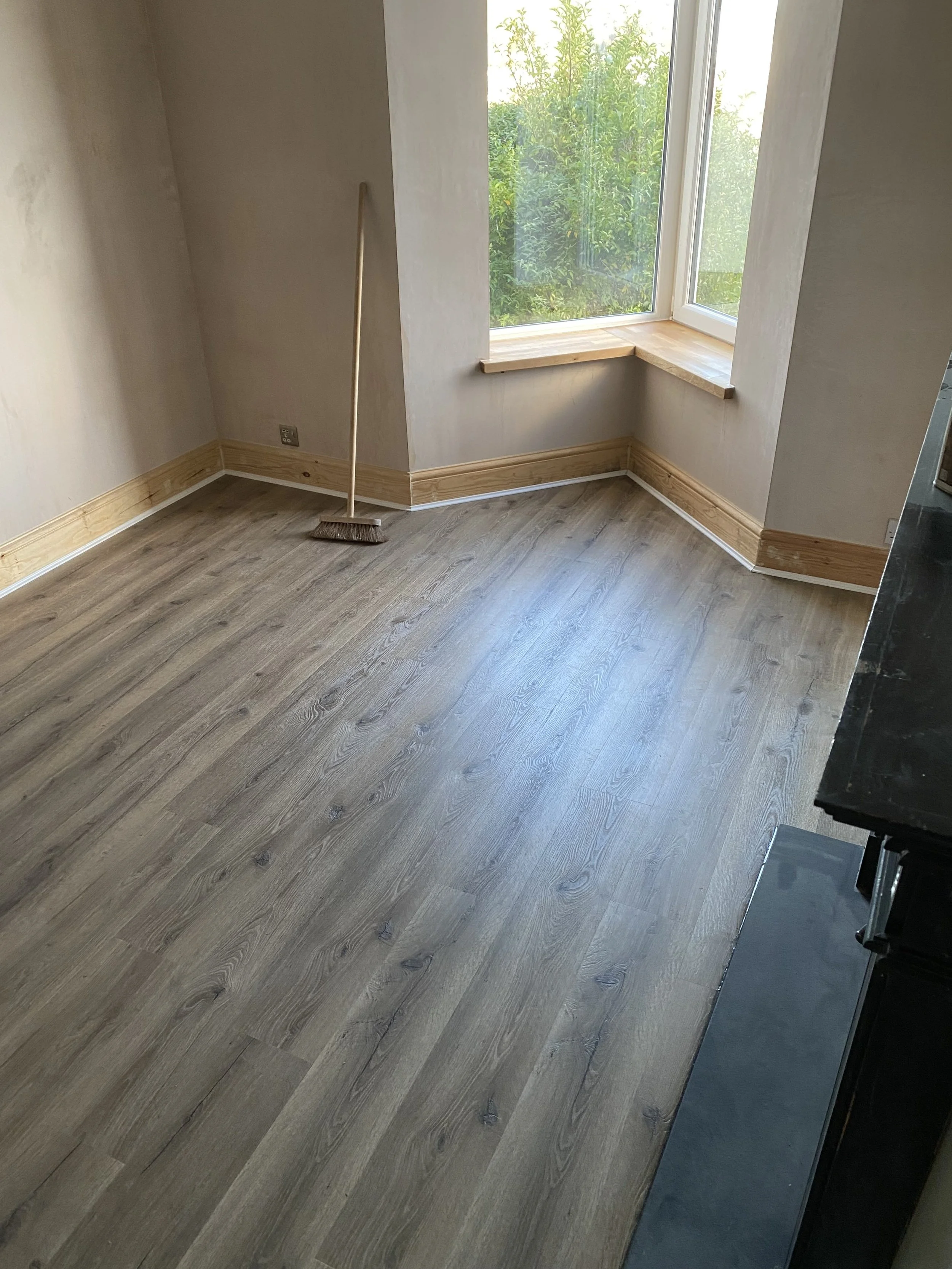Empty room with wood laminate flooring, large window with a wooden sill, and a broom leaning against the wall.