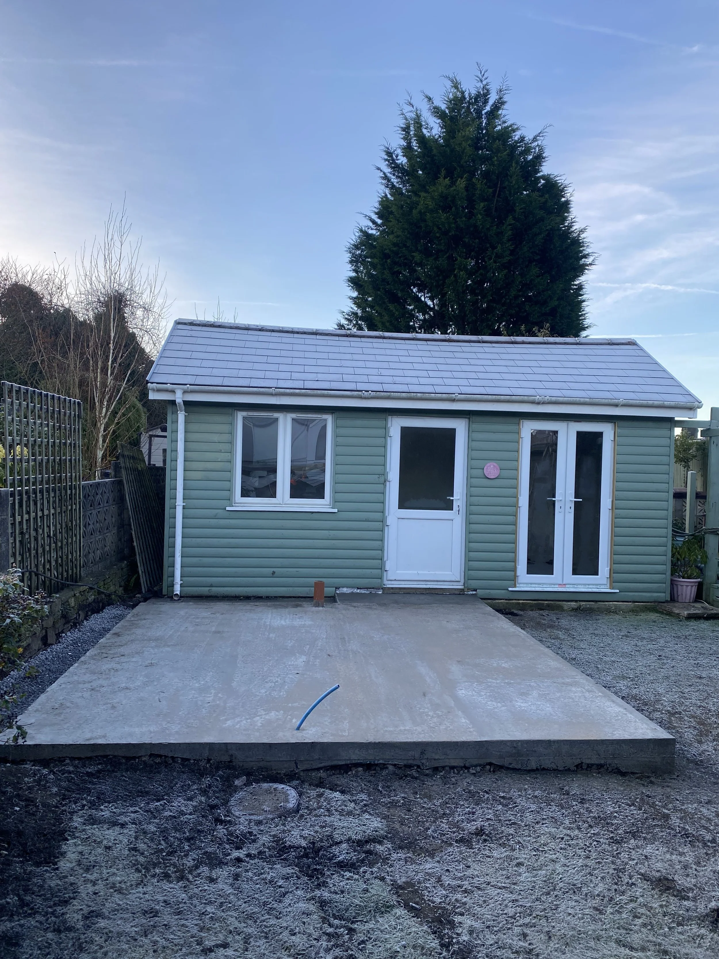 Small green vinyl-sided building with white trim, two doors on the right, a window on the left, and a large tree behind it, on a concrete pad with some frost, under a blue sky.