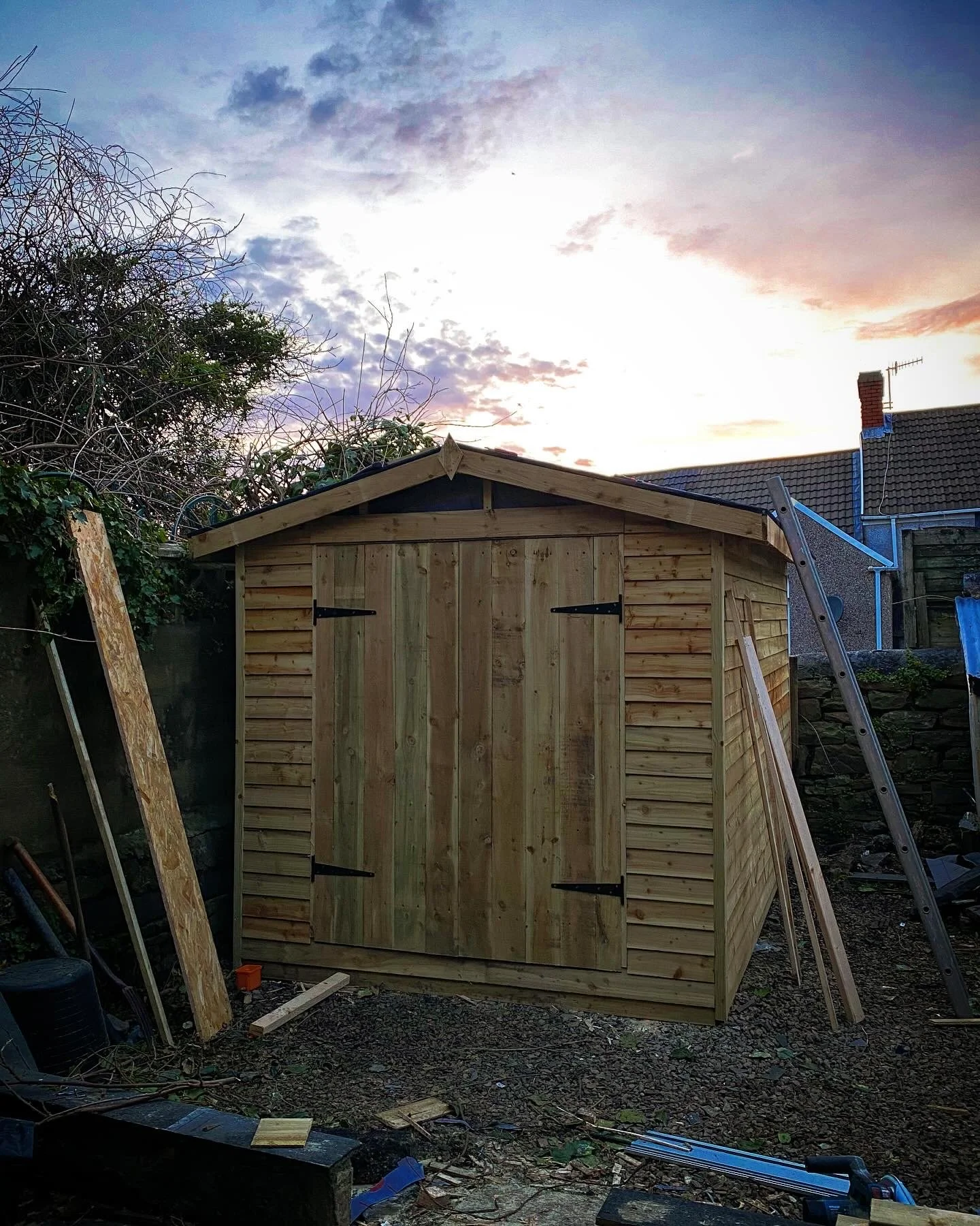 A newly built wooden shed with double doors, situated in a backyard during sunset, surrounded by tools and construction materials.