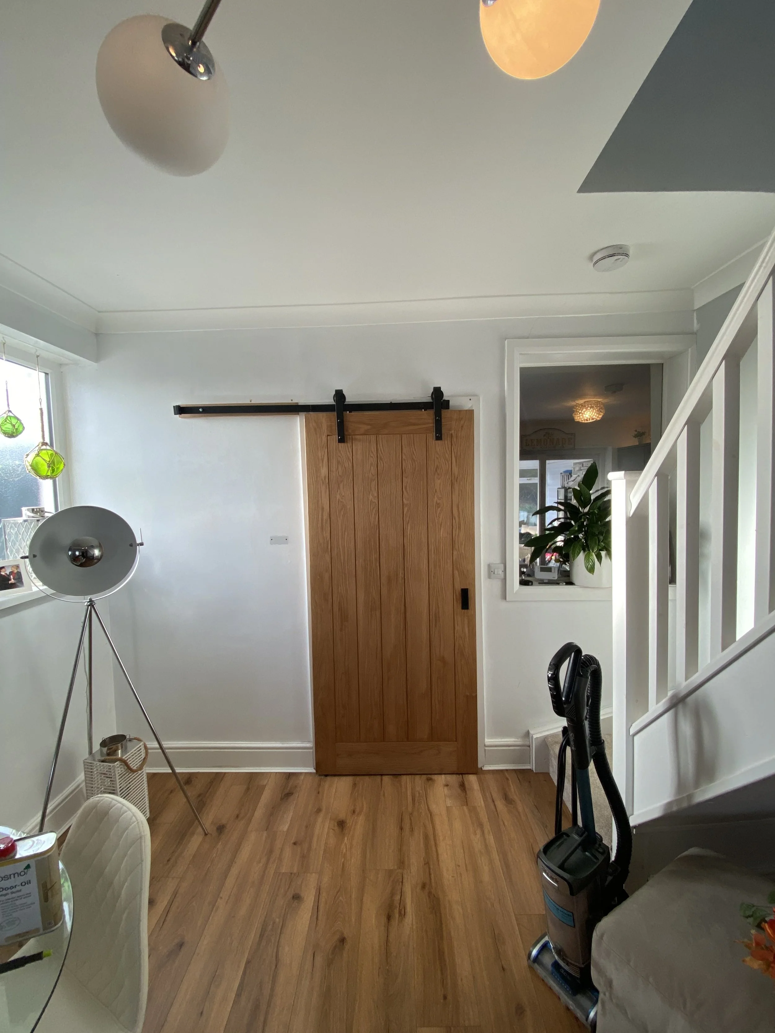 Interior view of a living room with a wooden sliding door, a window with green glass ornaments, a standing lamp, a plant in a white pot, a vacuum cleaner, and part of a staircase.