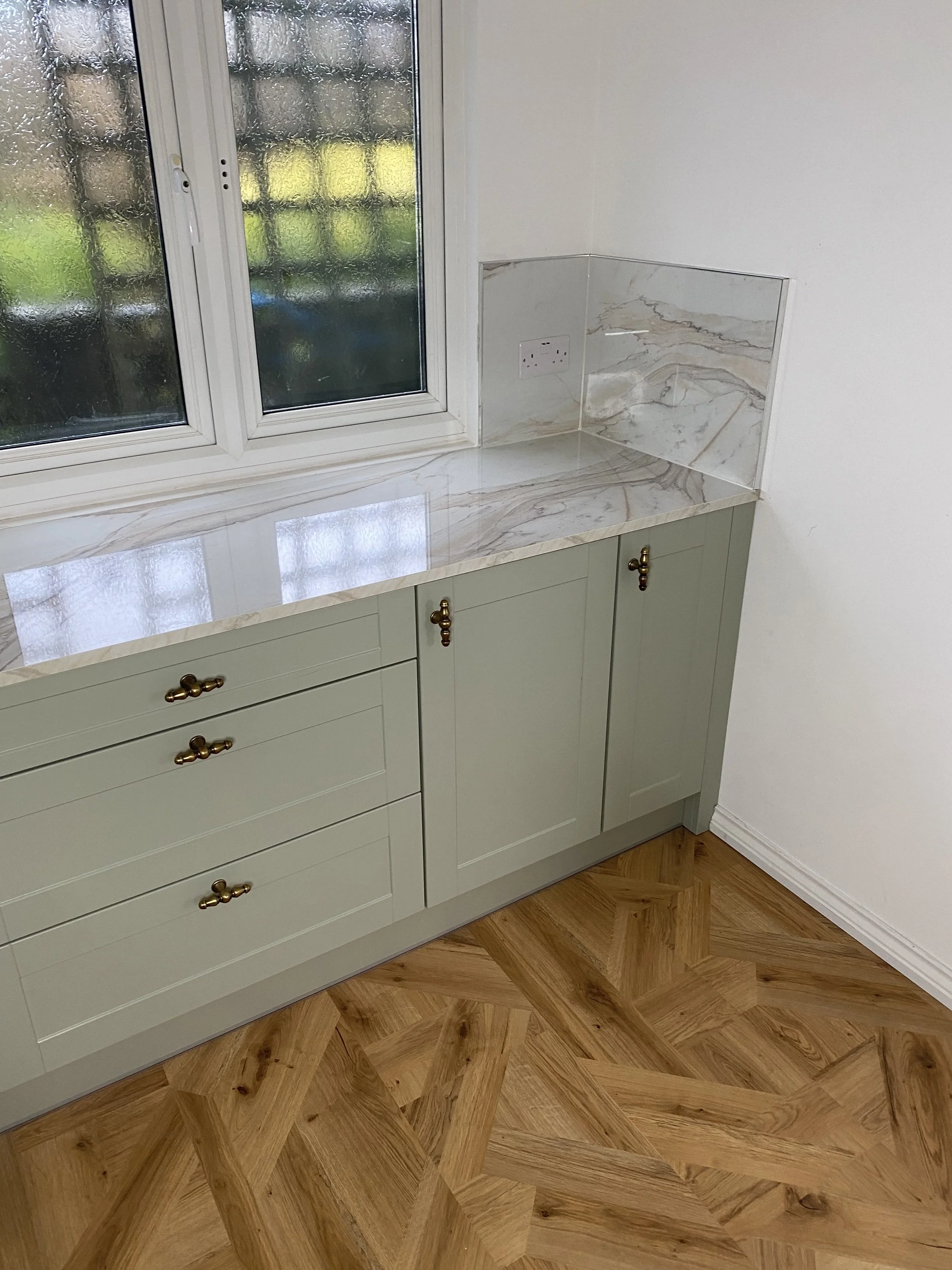A corner of a kitchen with a marble countertop, light green cabinets with bronze handles, a window with a rain-speckled glass pane, a white wall, and a wooden plank floor with a chevron pattern.