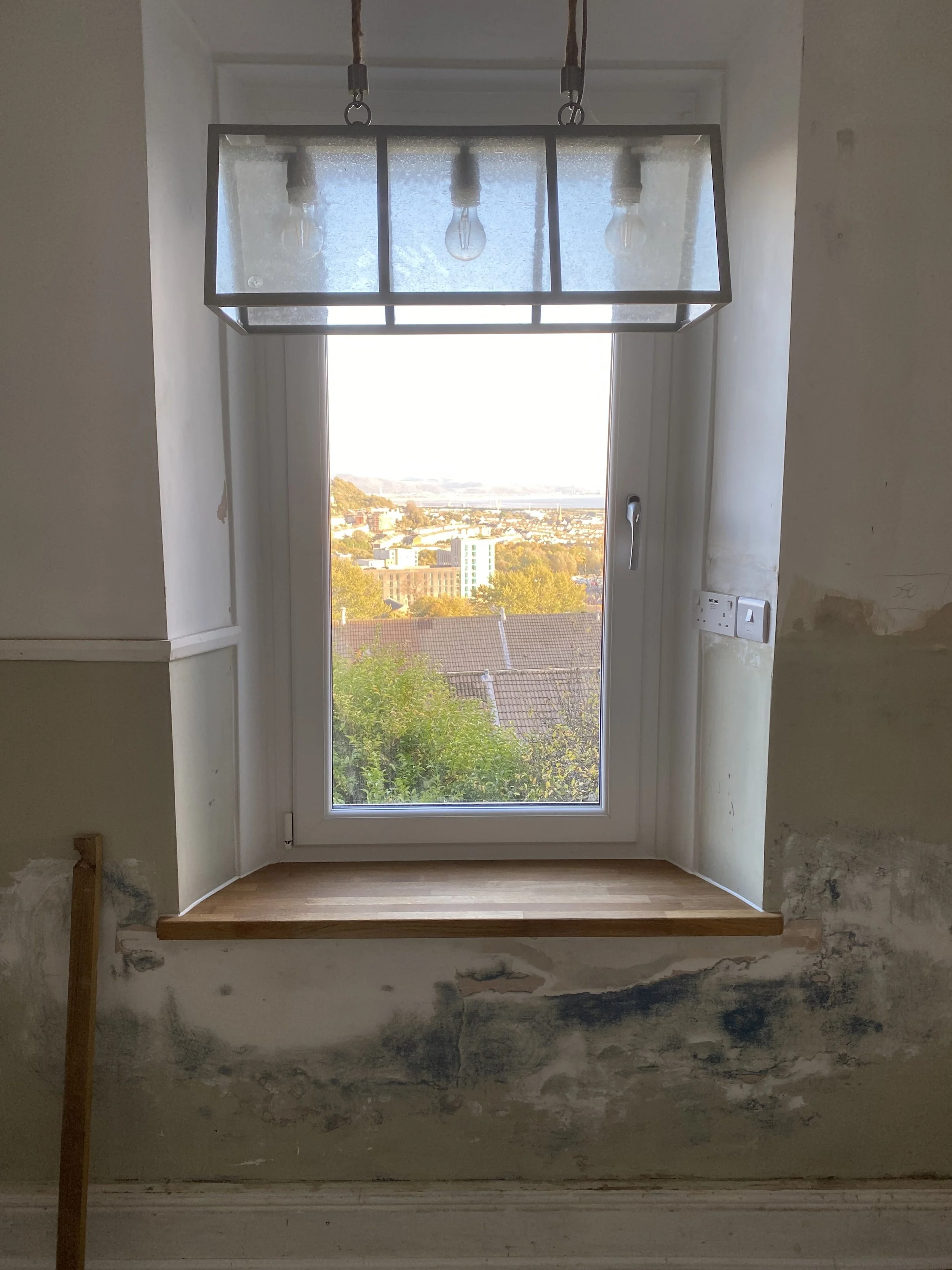 A window with a view of a cityscape and trees, with an unfinished wall and a wooden shelf beneath the window, and a modern rectangular hanging light fixture above.