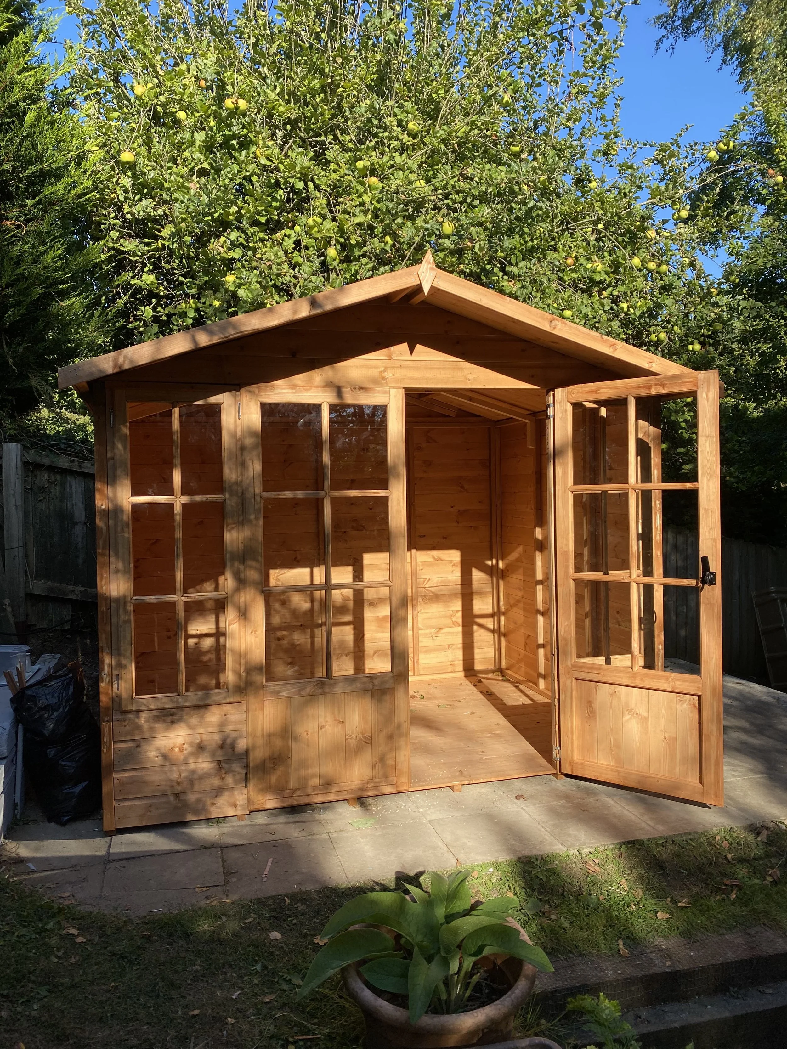A partially assembled wooden garden shed with open doors, positioned on a stone patio, with a green tree full of yellow apples in the background, and a potted green plant in the foreground.