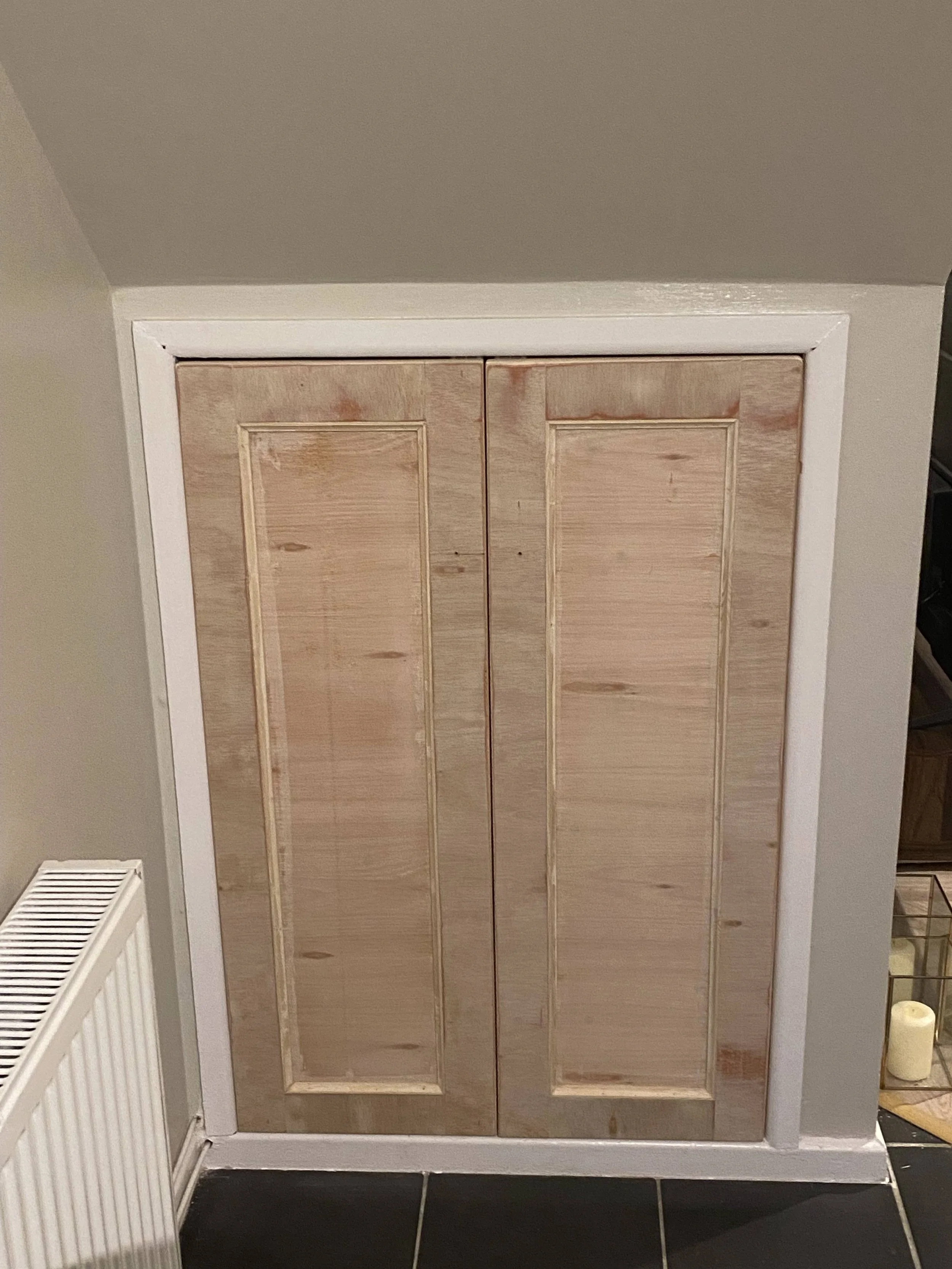 Unfinished wooden cabinet with vertical panels, framed by white trim, located in a room with taupe walls and black tiled flooring.