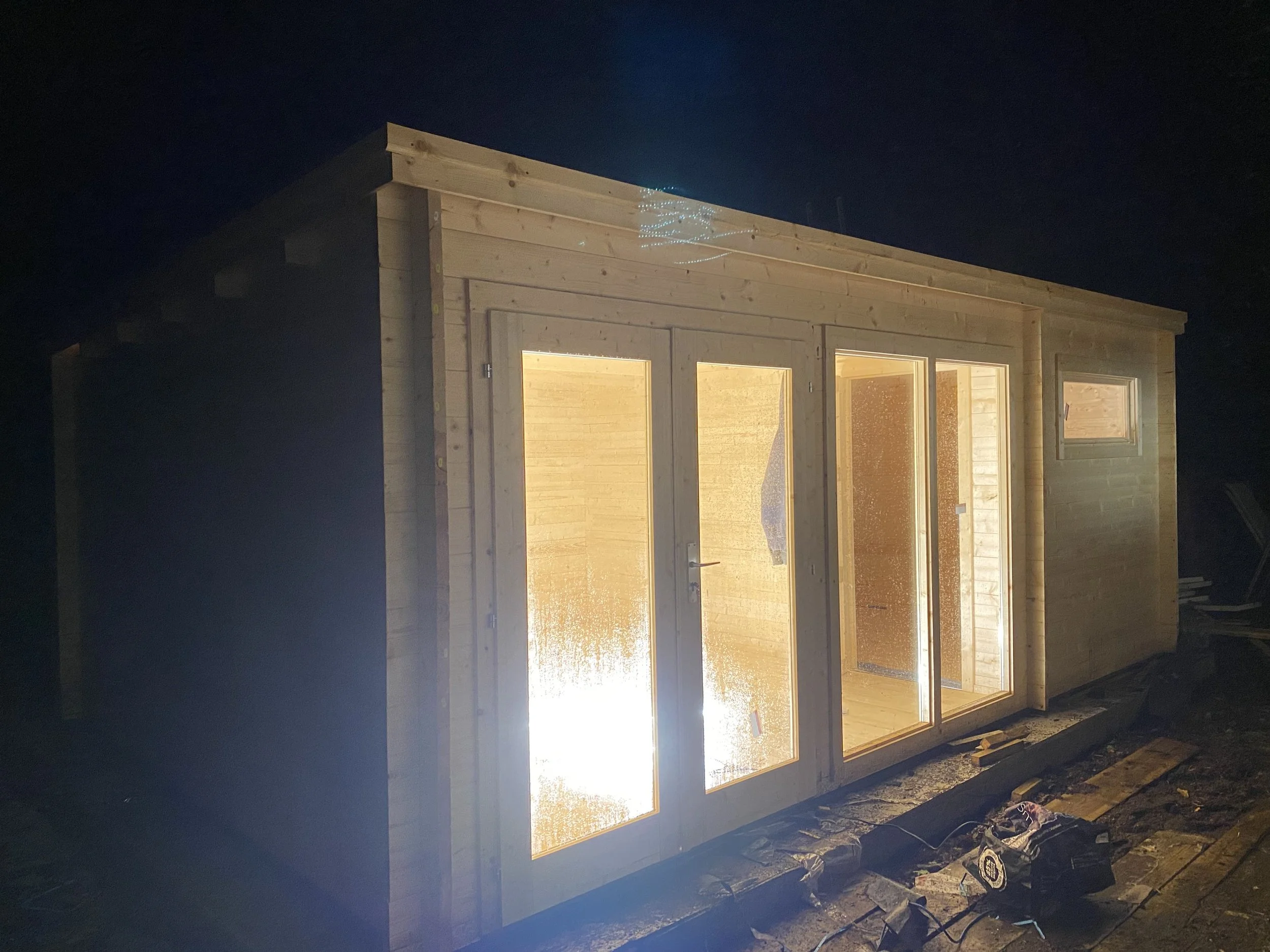 Wooden shed under construction at night, with interior lights on, showing double glass doors and a small window, surrounded by construction materials.