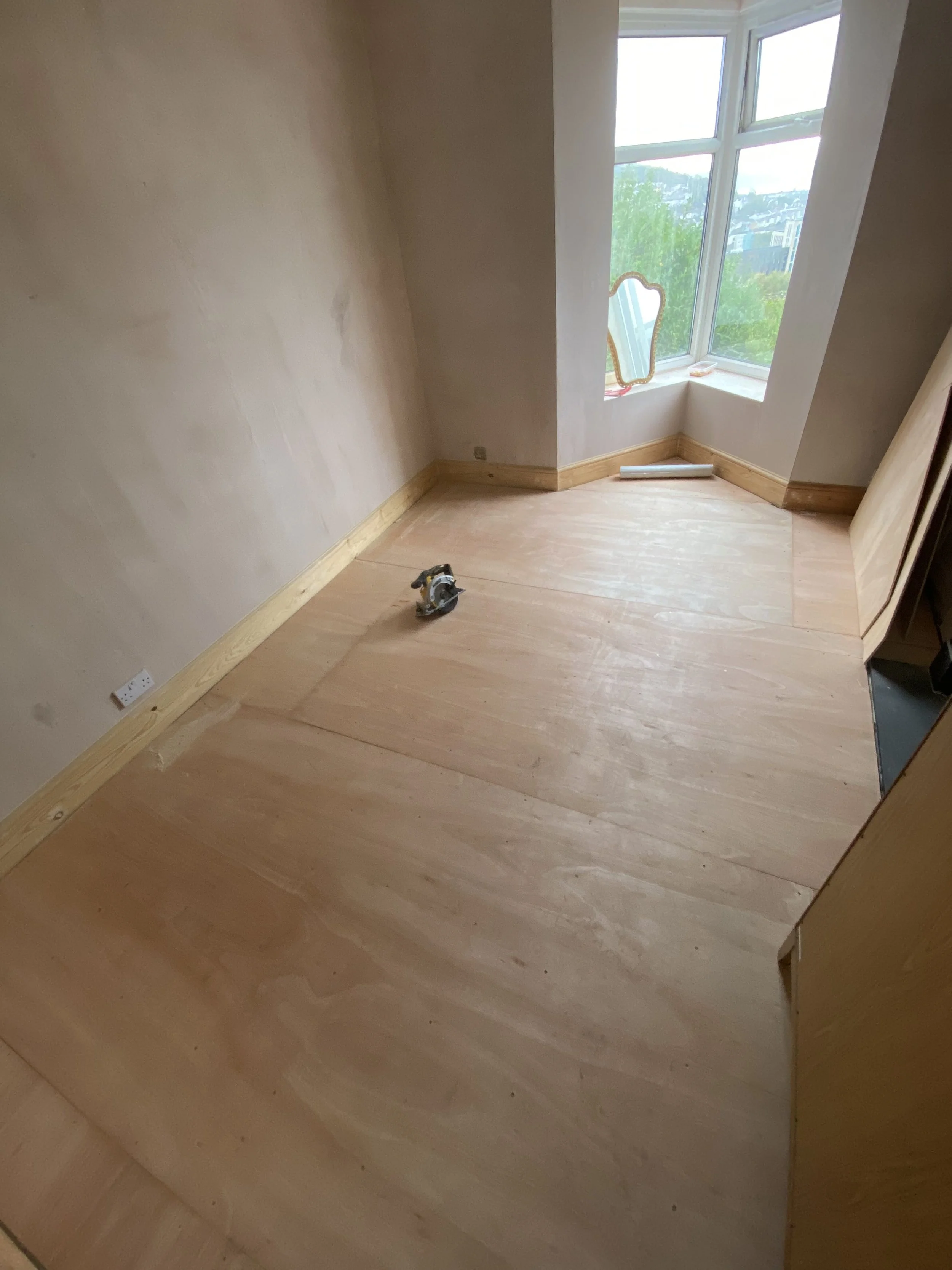 Room under renovation with wooden flooring partially installed, a power tool on the floor, an angled window with a reflection mirror, and a view of a neighborhood outside.