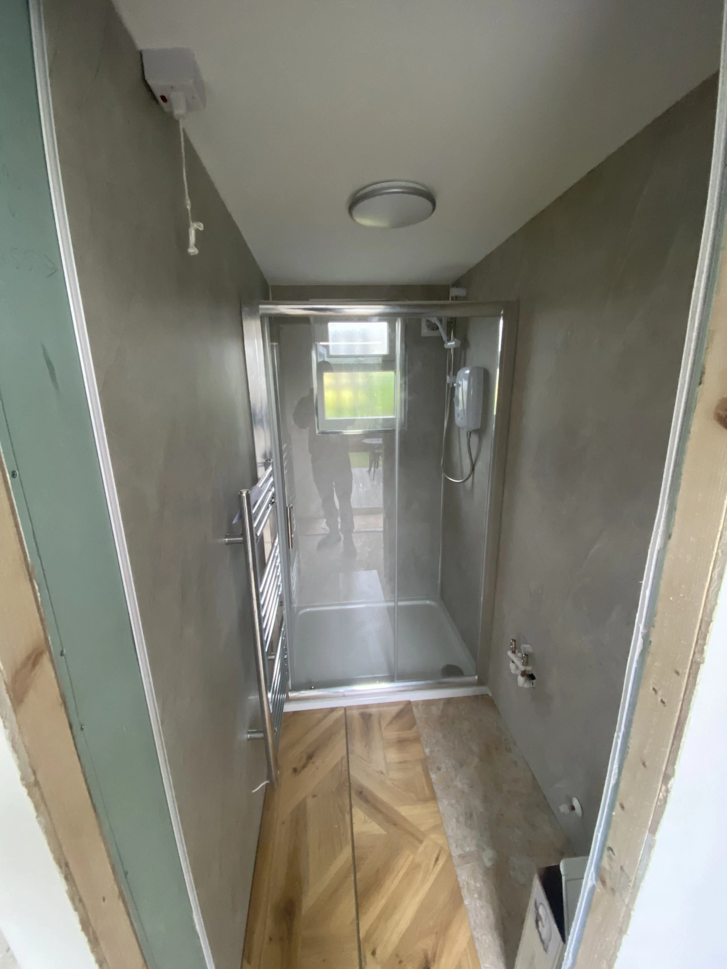 Small bathroom with a glass shower stall, a small window, and unfinished walls. A towel radiator is mounted on the left wall, and a ceiling light fixture is present. The bathroom has wooden and concrete flooring.