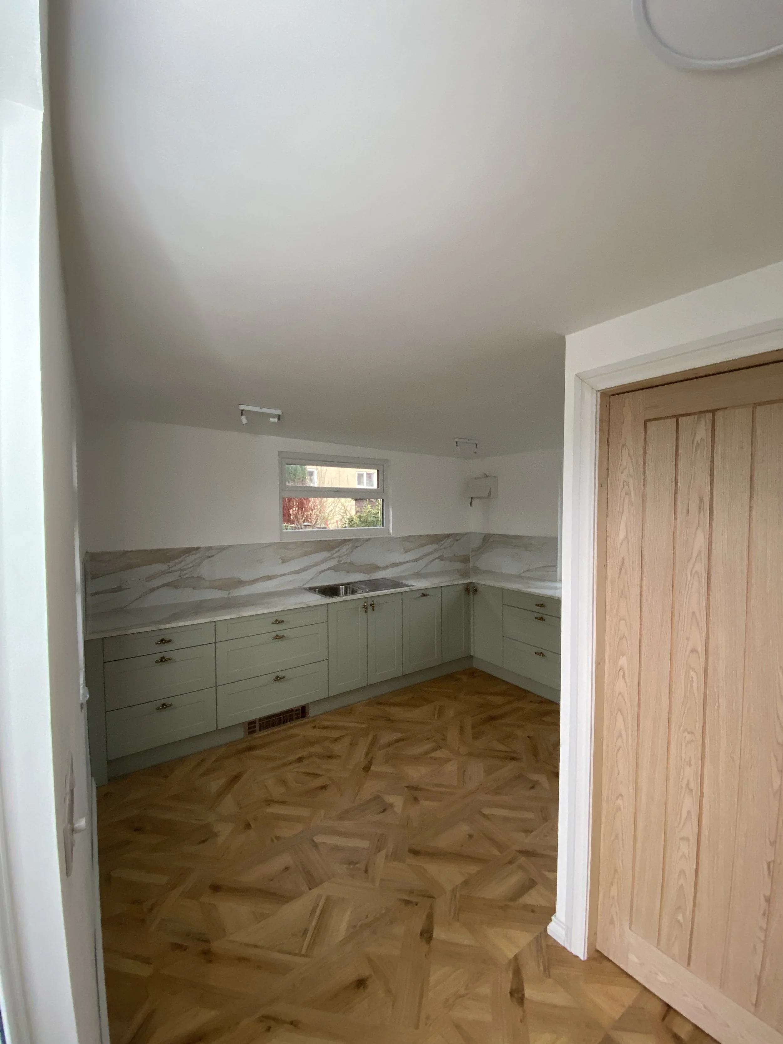 The image shows a kitchen with light green cabinets, a marble backsplash, a small window, a wooden floor, and a wooden door.