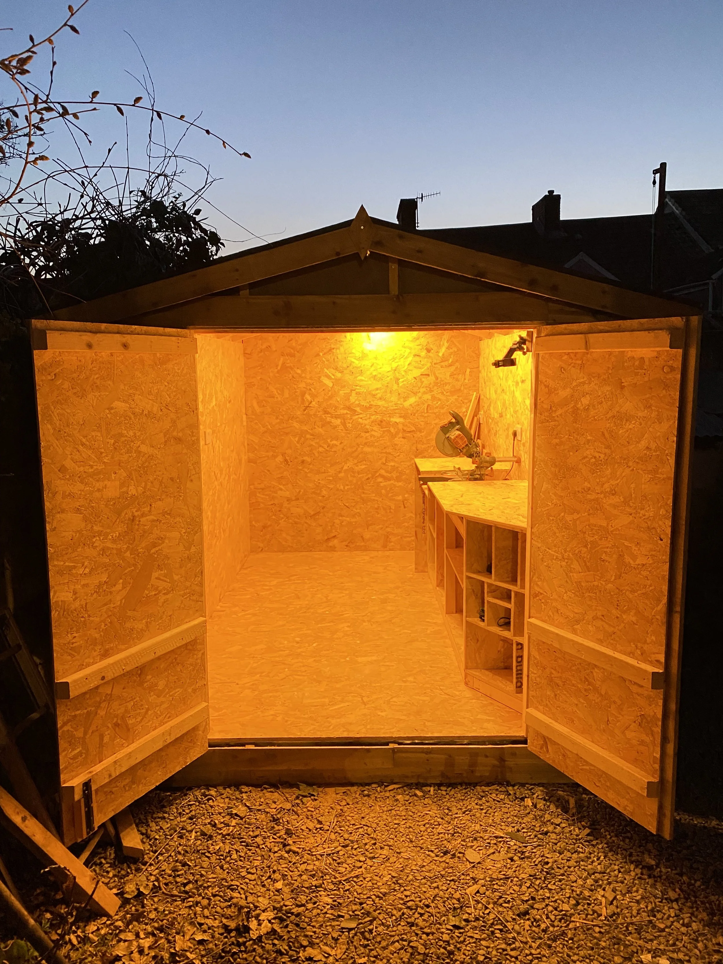 Inside a small wooden shed with unfinished plywood walls and floor, illuminated by a bright yellow light, with a workbench and power tools visible on the right side.