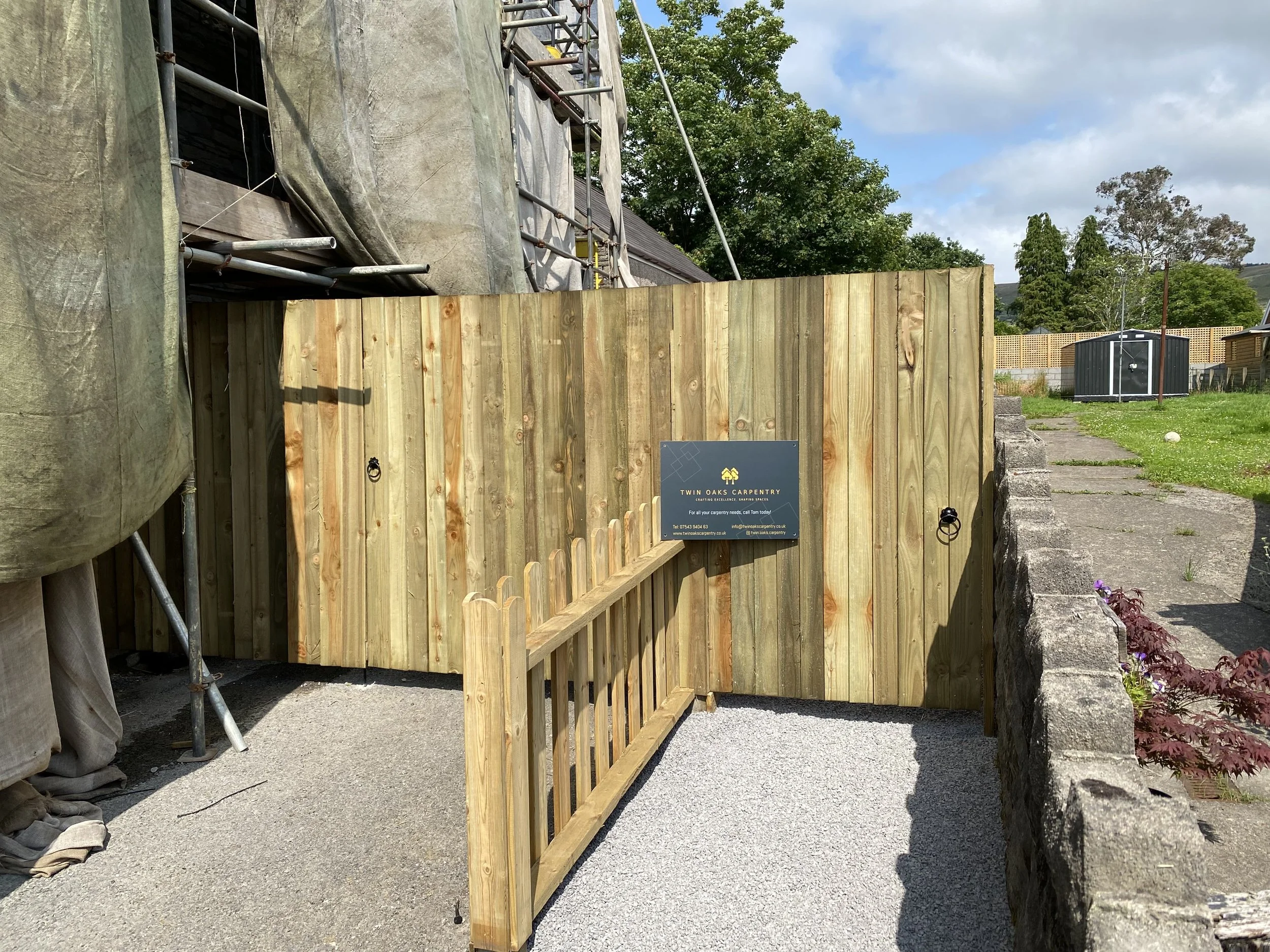 A newly constructed wooden fence with a small gate, located outdoors on a gravel surface, with a sign reading 'Twin Oaks Carpentry' attached to the fence.