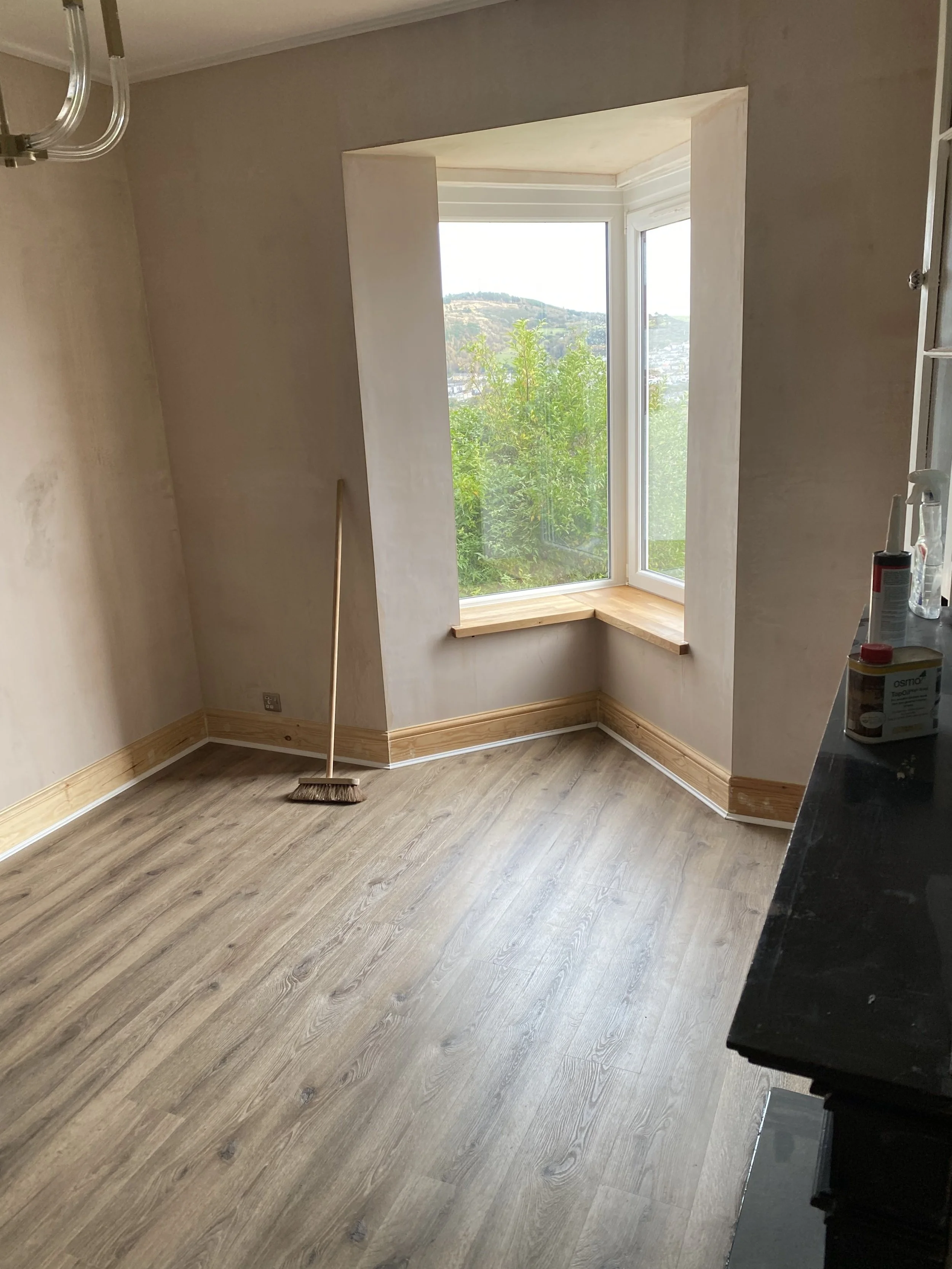 Empty room with wooden flooring, partially painted walls, and a large bay window showing green trees and hills outside, with a broom leaning against the wall.