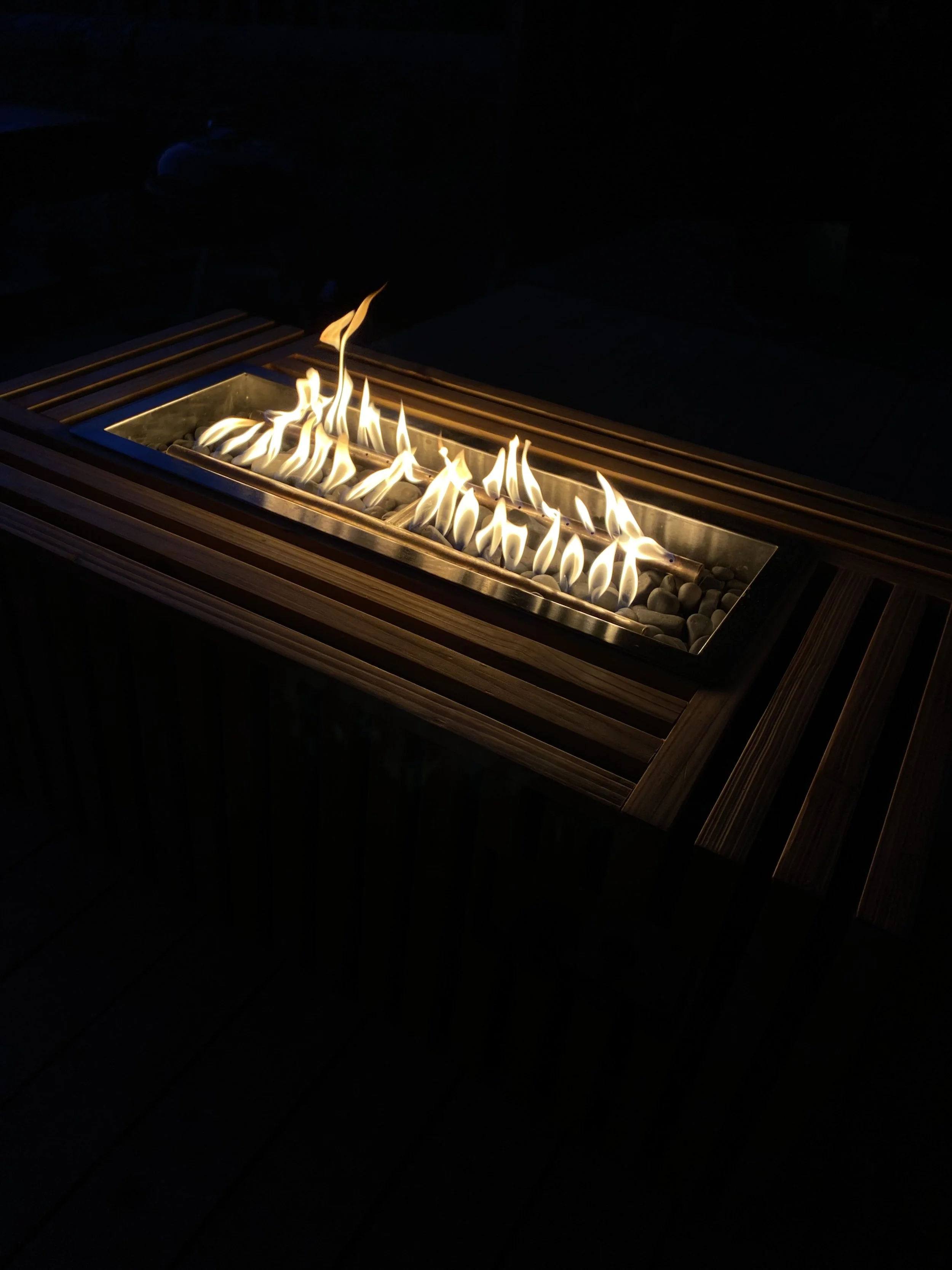 Outdoor fire pit with flames burning on a bed of rocks, surrounded by a wooden frame and slats, in darkness.