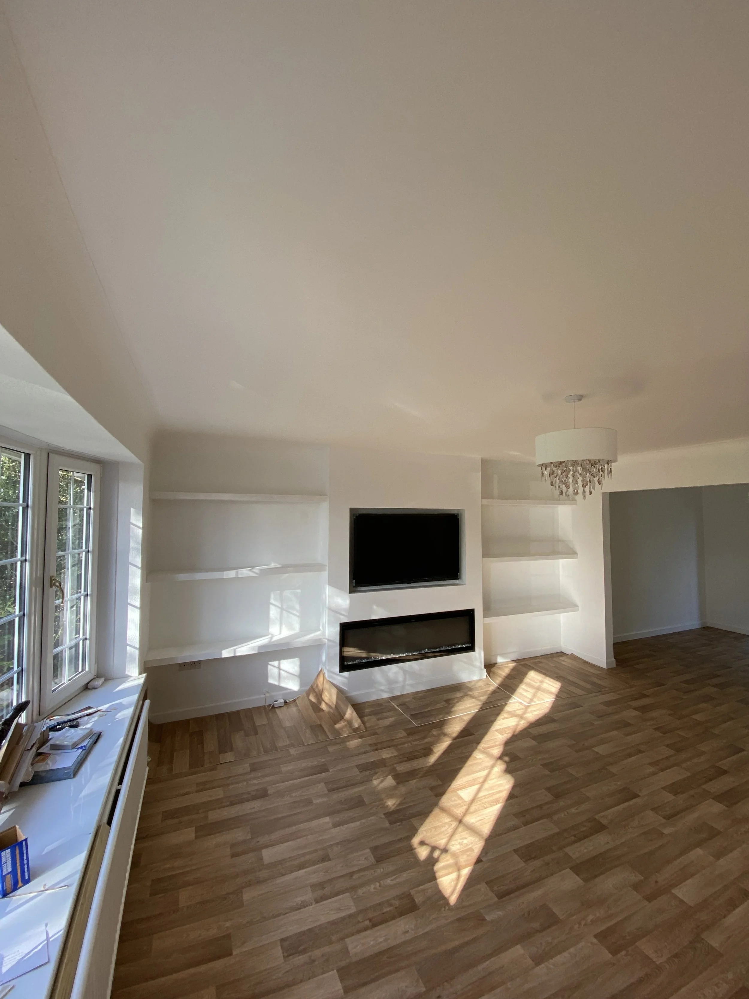 Empty living room with hardwood floors, white walls, built-in shelves, a mounted TV and fireplace, a large window, and a chandelier.