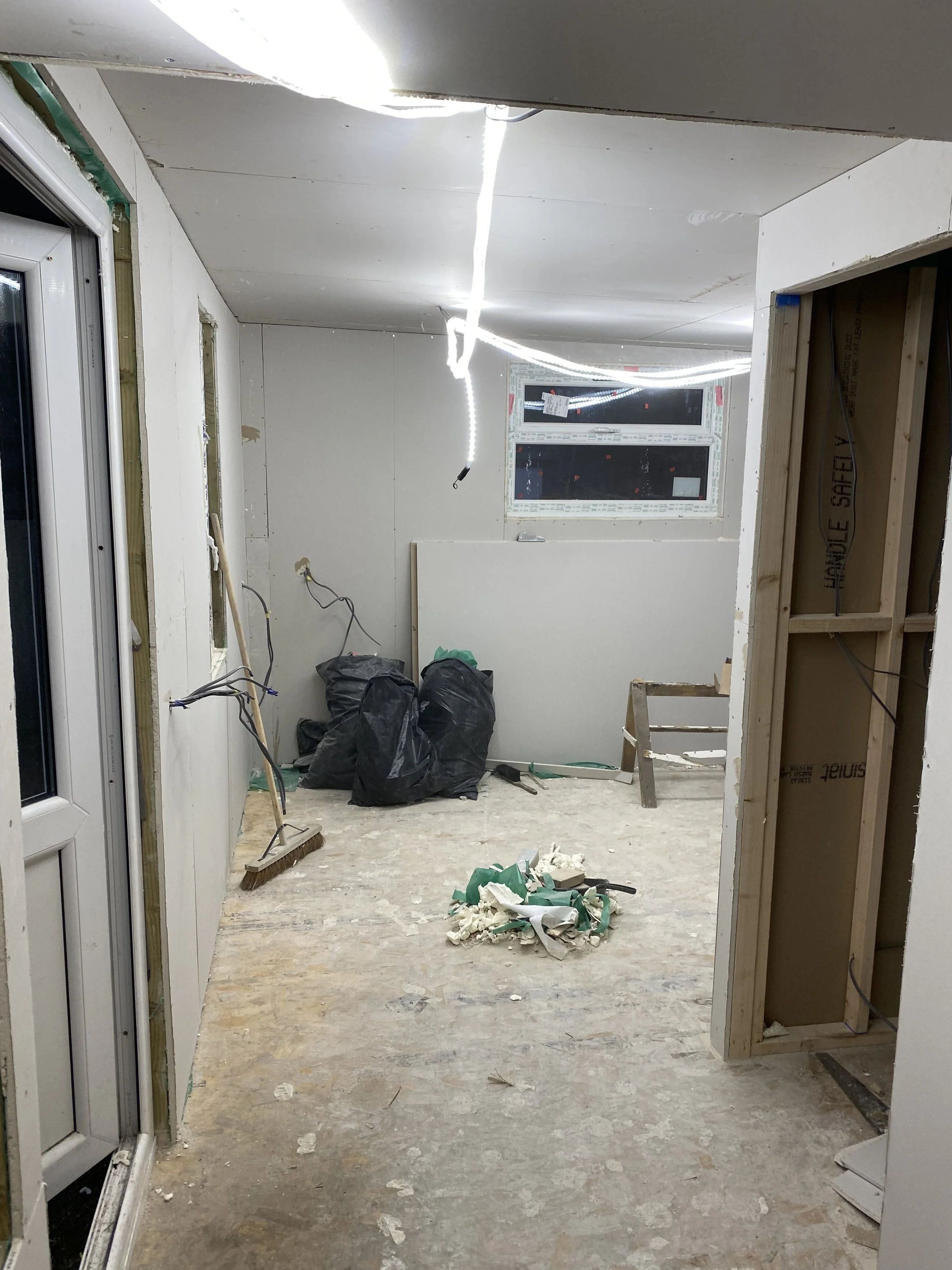 Room under renovation with unfinished walls, construction debris on the floor, a broom leaning against the wall, black trash bags in the corner, and exposed electrical wiring.
