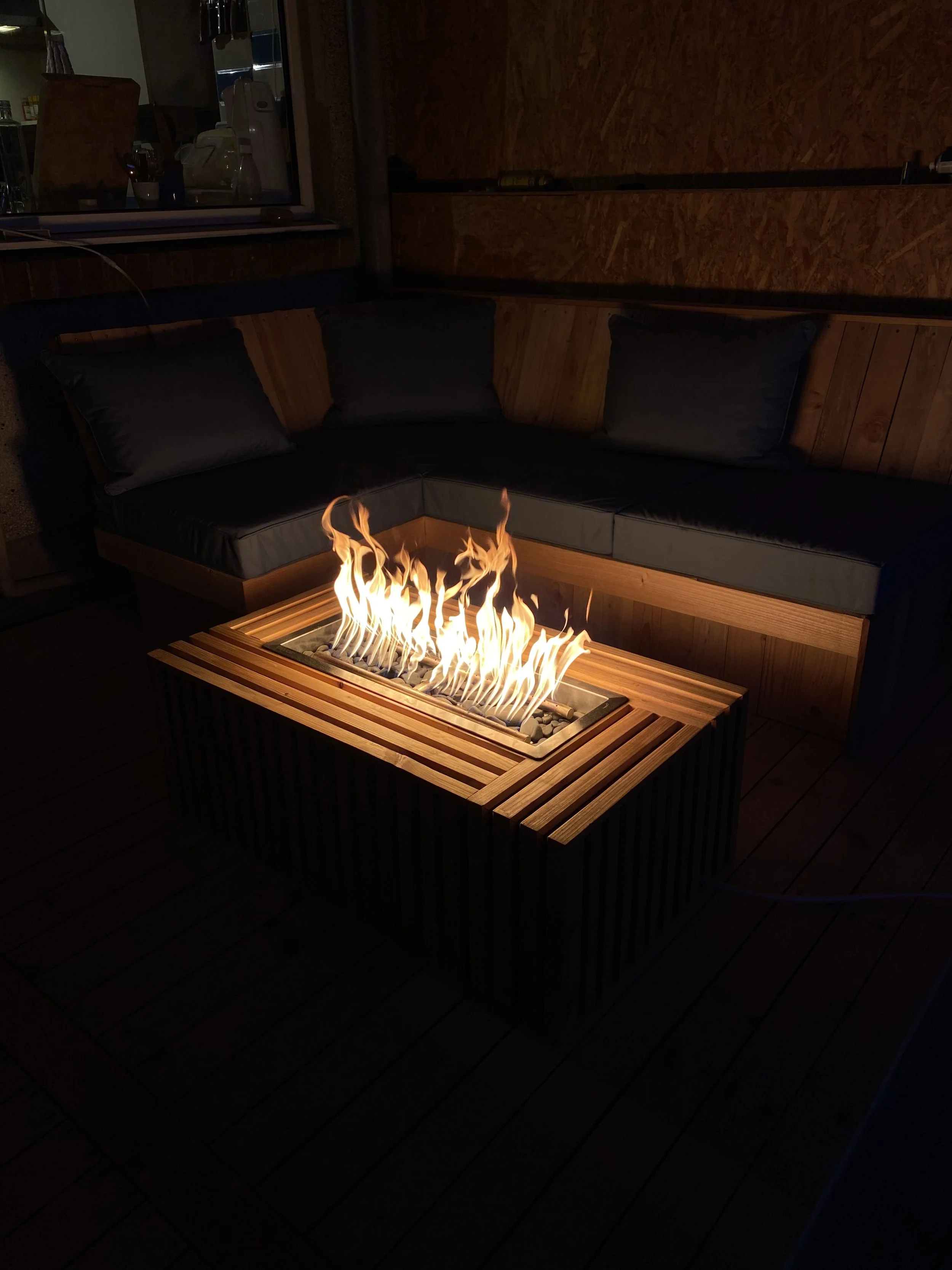 Outdoor patio with a modern fire pit table featuring flames, surrounded by a sectional sofa with black cushions, in a dimly lit setting.