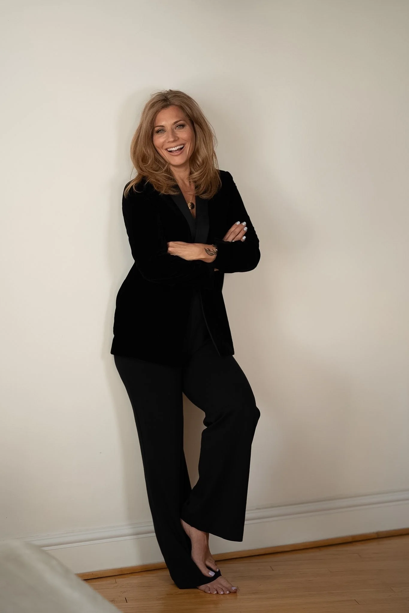 A woman in black attire with blonde hair standing against a plain wall, smiling with crossed arms.