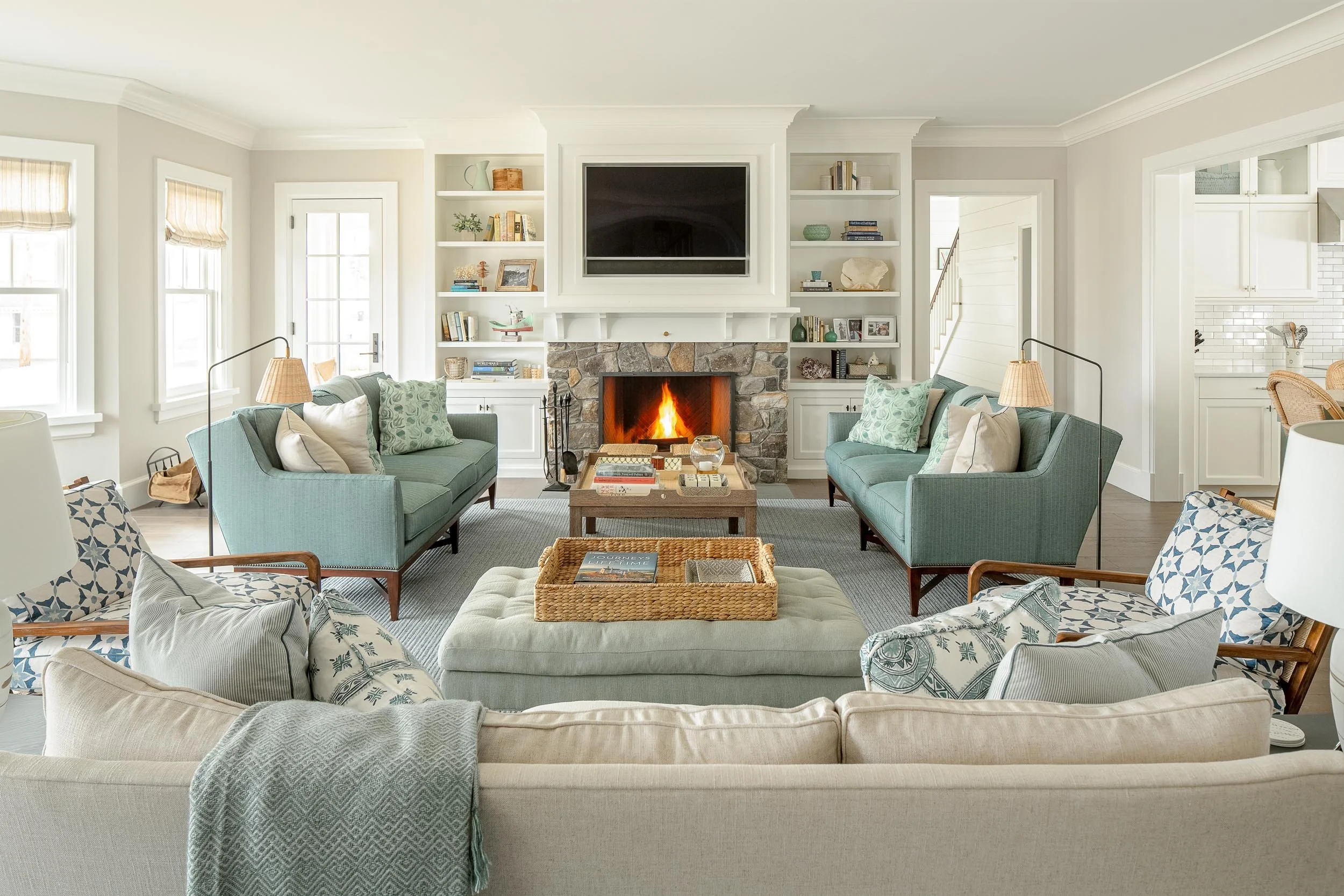 Living room with a fireplace, built-in white shelves, two green sofas, a beige armchair with a patterned pillow, a coffee table with books, a woven tray, and a fireplace with a fire. Windows with beige Roman shades and a doorway visible.