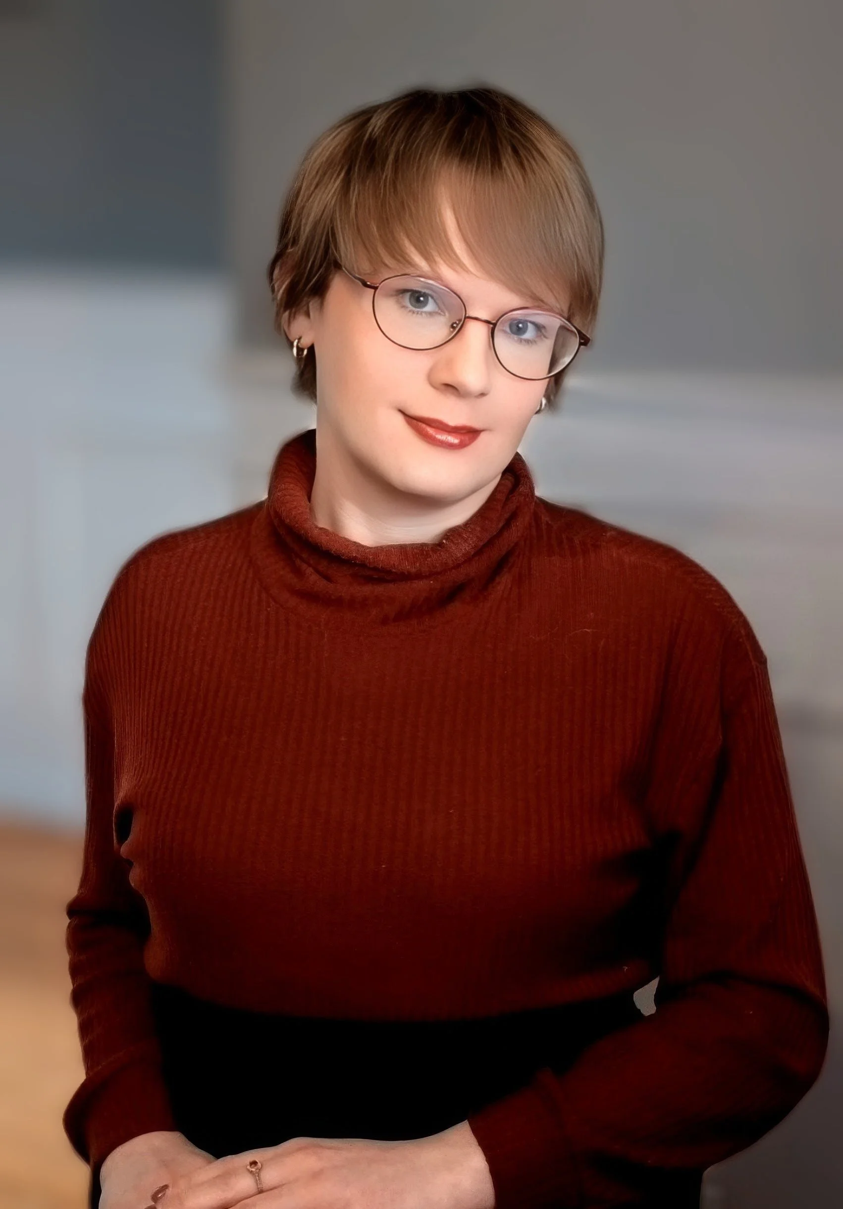 A woman with short light brown hair wearing glasses, a red turtleneck sweater, and jewelry, sitting indoors with a blurred background.