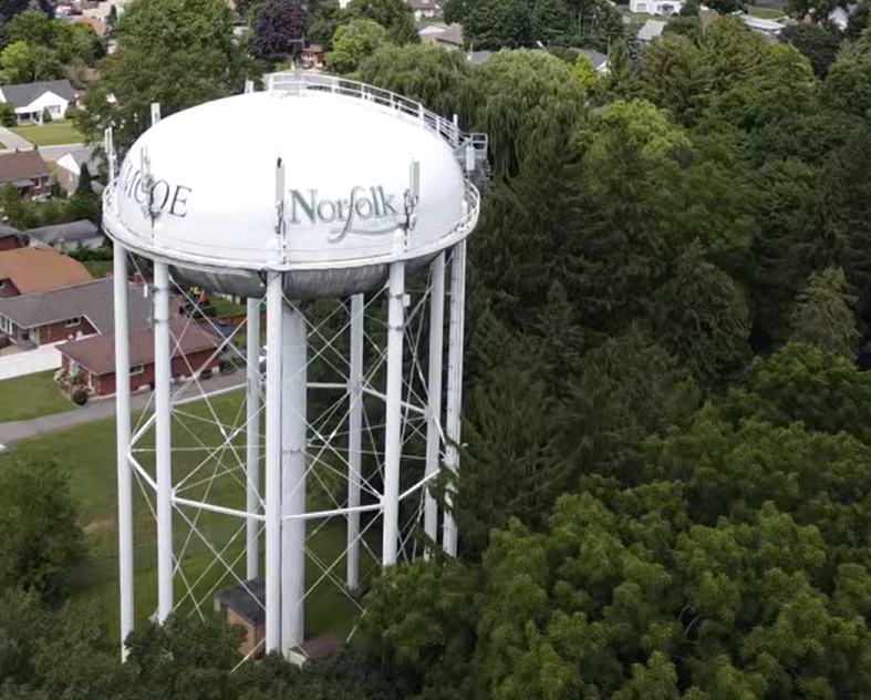 An aerial view of a white water tower with the words 'Norfolk'  visible, surrounded by green trees and residential houses.