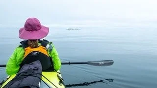 Person kayaking on calm water, wearing a pink hat and green jacket, with distant islands on the horizon.