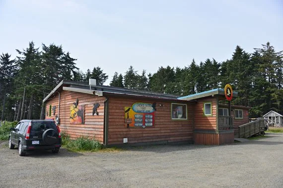 A small wooden restaurant or diner with colorful signs on the exterior wall, located in a forested area with trees in the background. A black SUV is parked in front of the building.