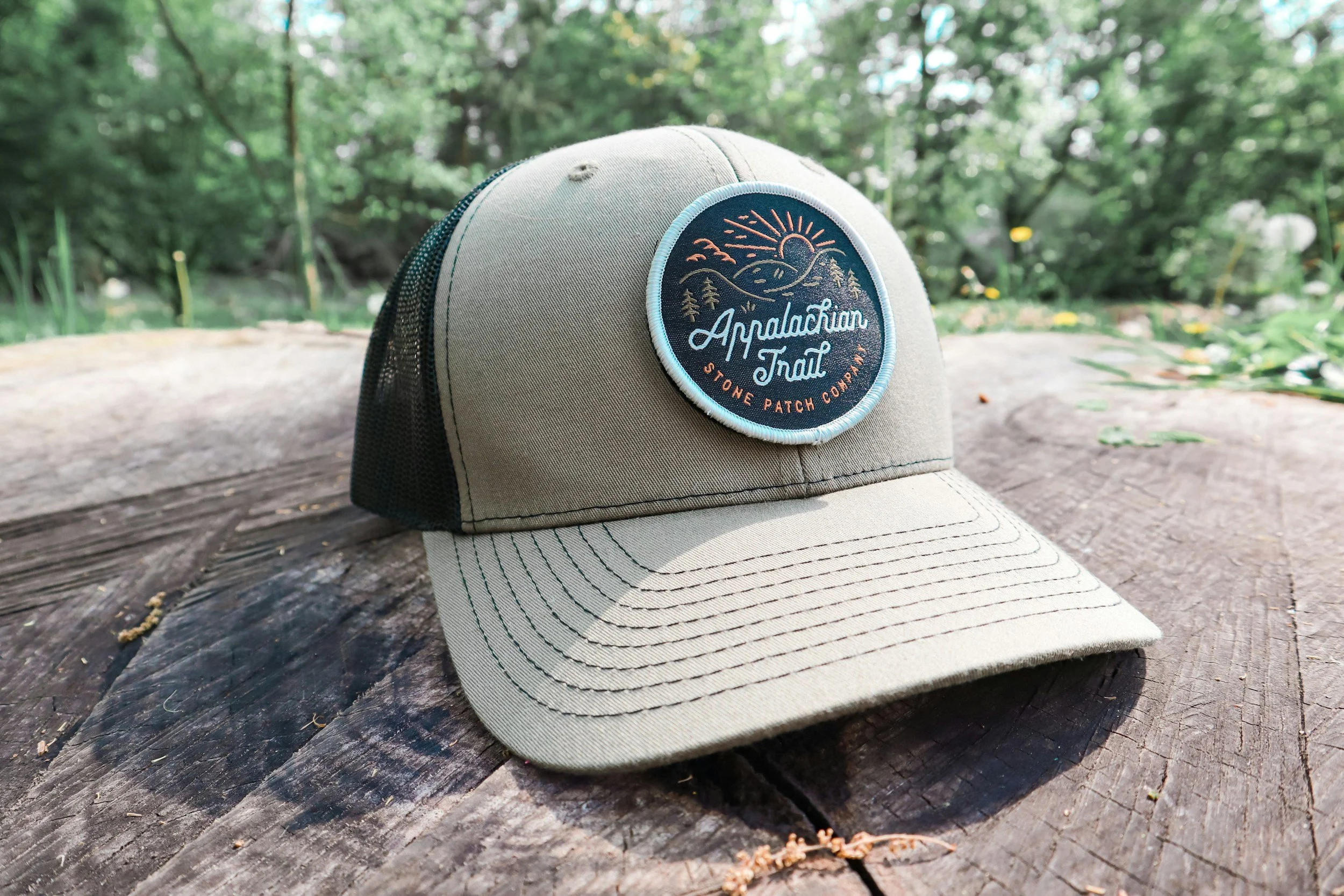 A beige and black mesh trucker hat with an embroidered patch on the front, featuring a mountain scene, a sun, and the words 'Appalachian Trail, Stone Patch Company', resting on a weathered wooden surface outdoors surrounded by greenery.