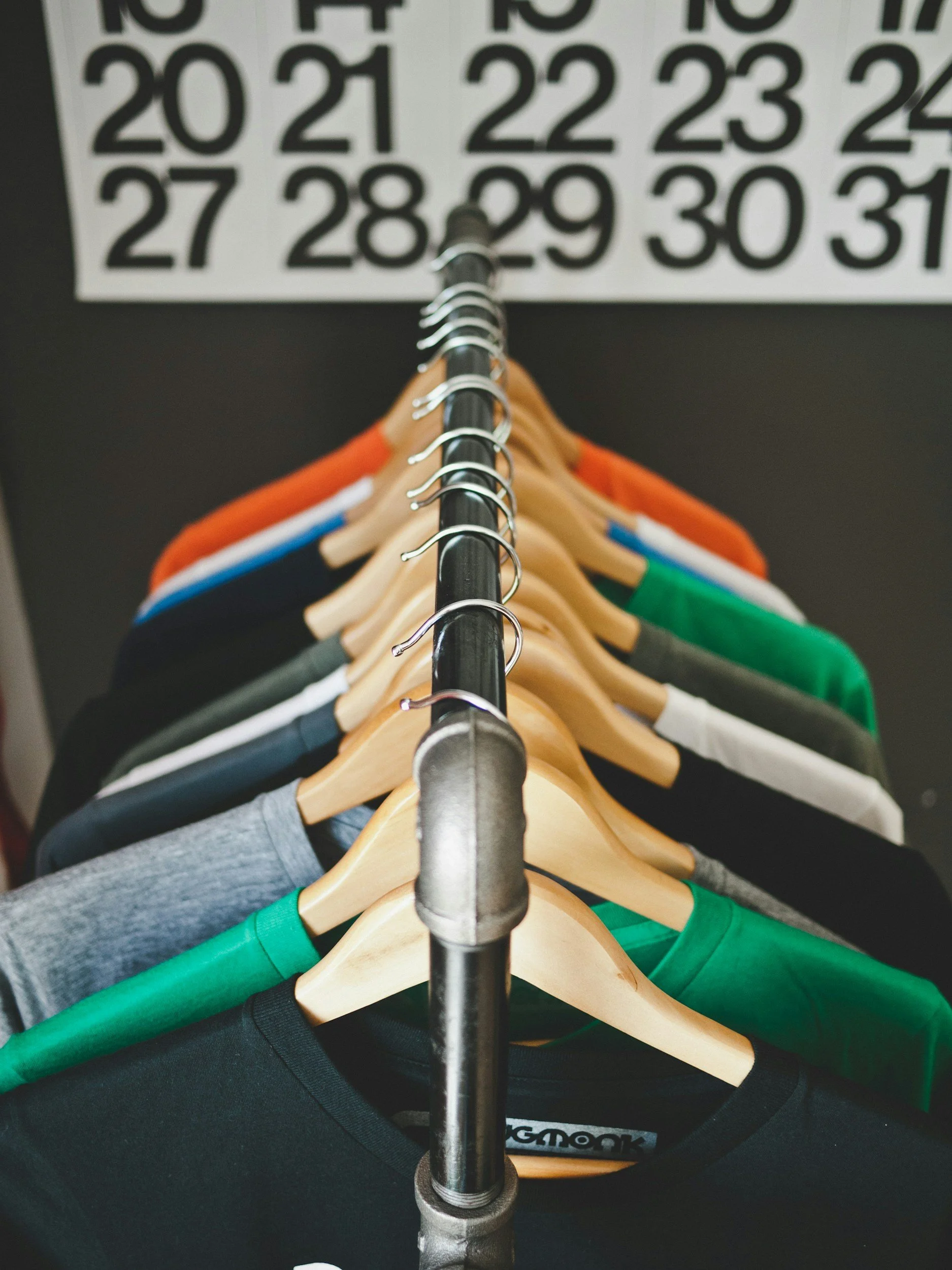 A row of assorted T-shirts on hangers hanging on a metal rod in front of a wall calendar showing dates from 20 to 31.