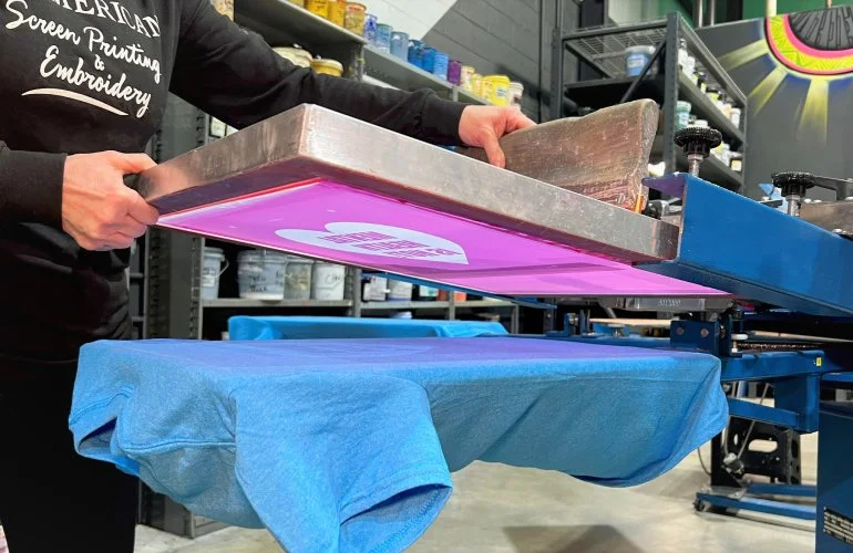 Person operating a flatbed screen printing press with a pink light, printing on a blue t-shirt.