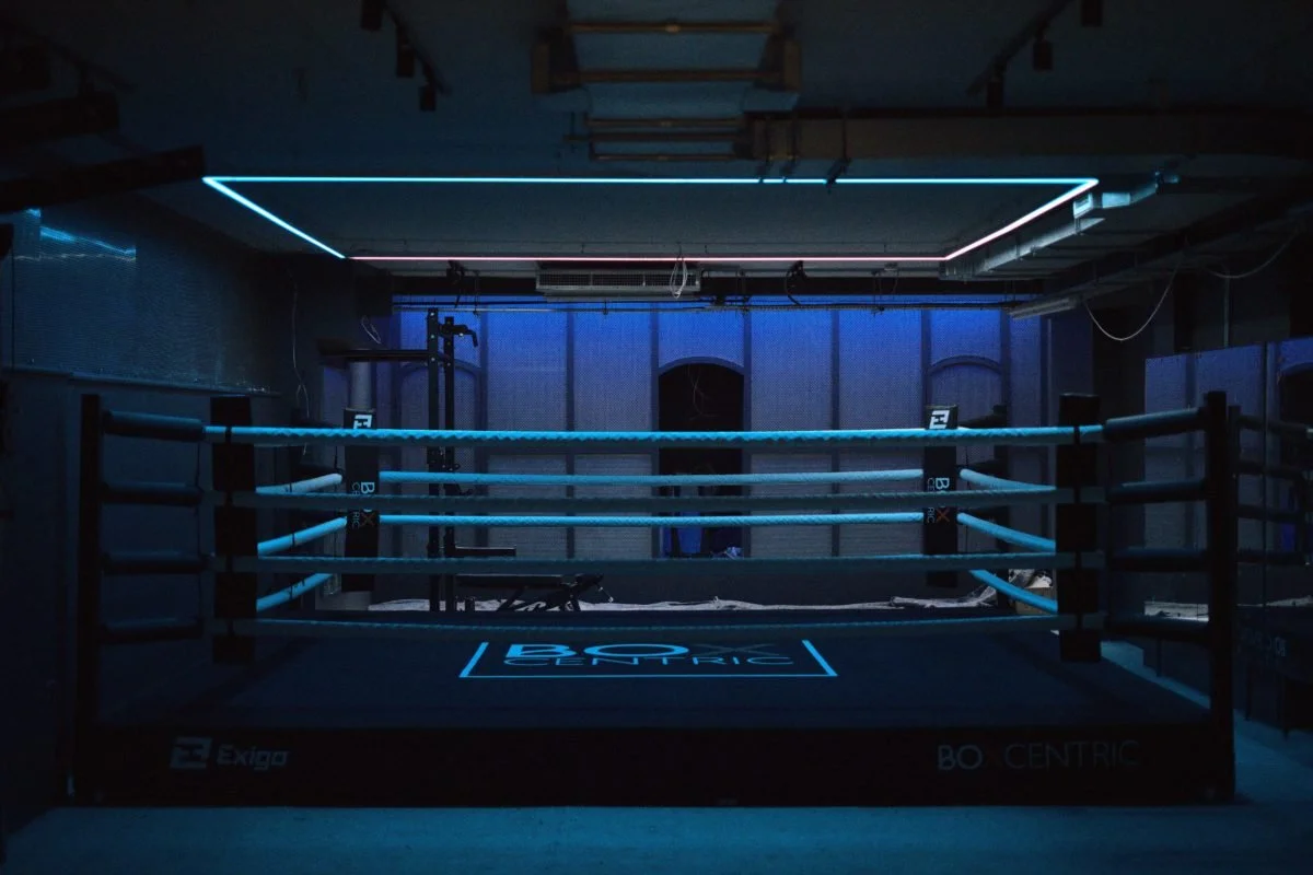 Dark boxing ring with illuminated blue and pink neon lights in a dimly lit gym.